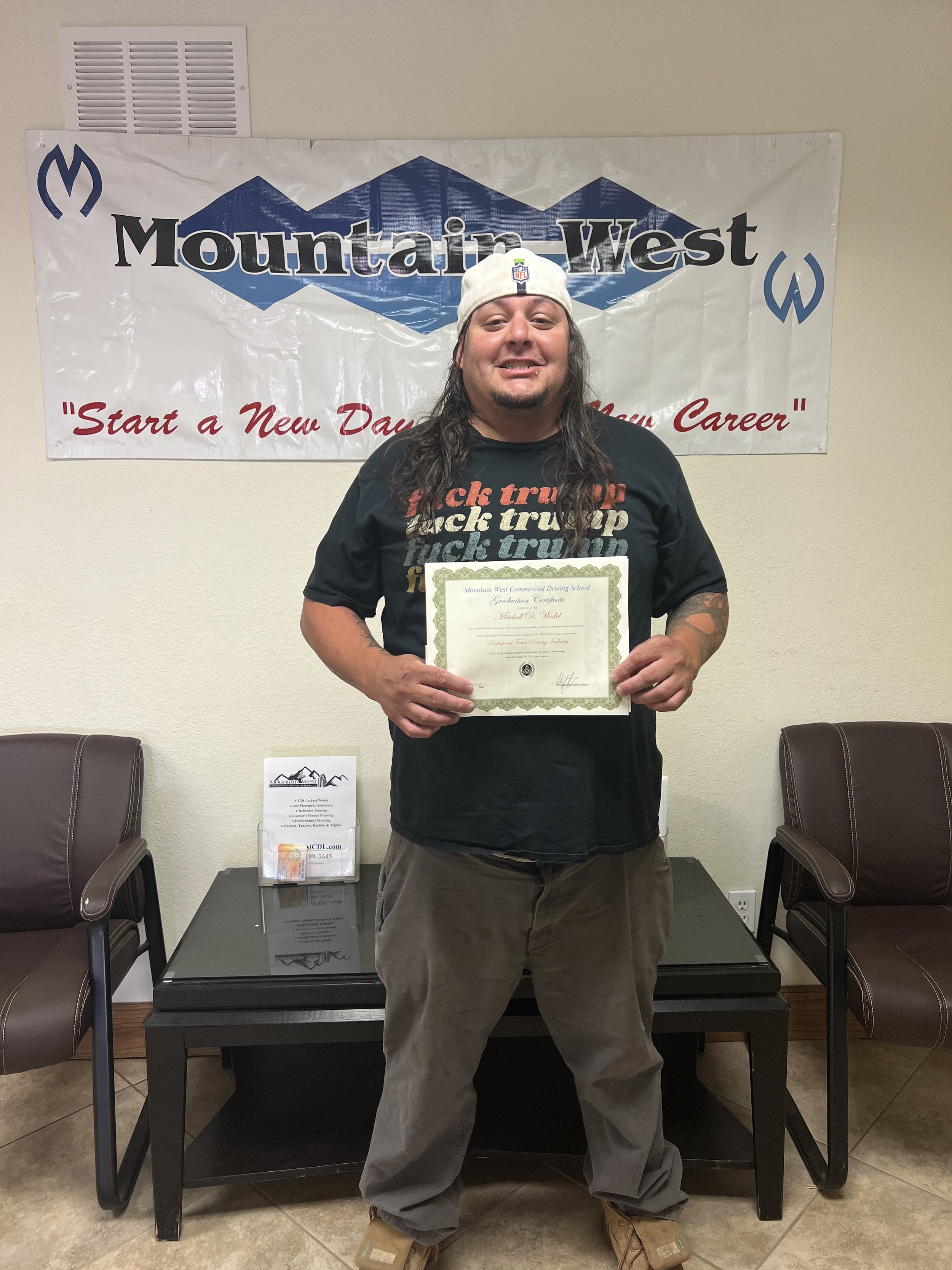 A man with long hair and a beard, wearing a white baseball cap and black t-shirt, standing in front of a Mountain West banner, holding a certificate.