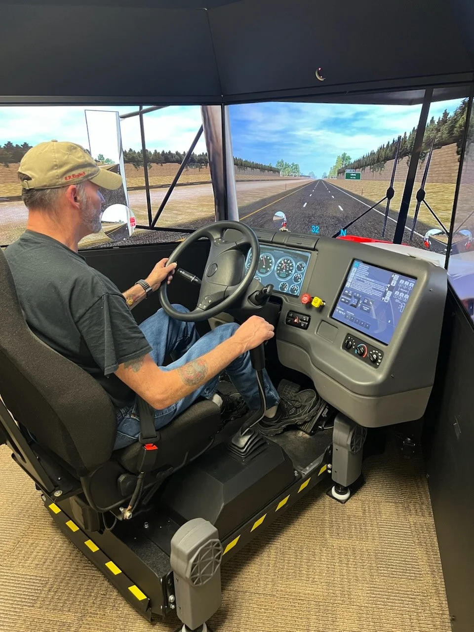 Man holding a steering wheel inside a truck simulator with digital dashboard and a large screen displaying a road scene.