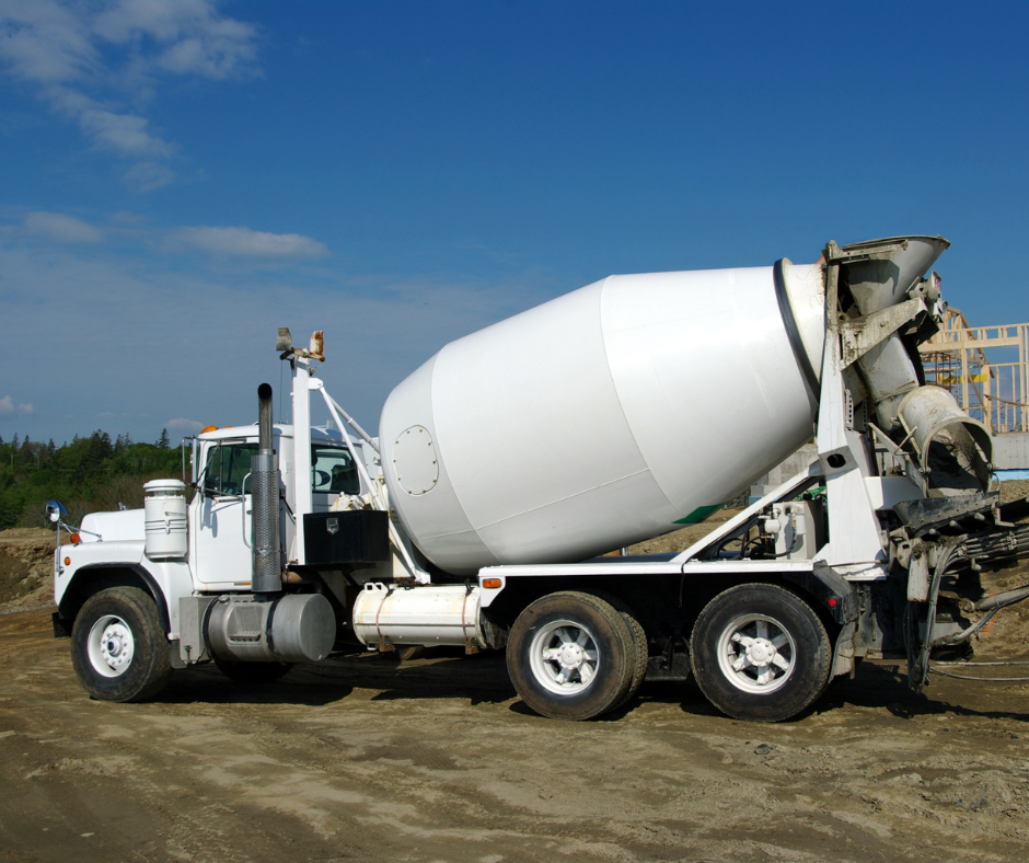 A white cement mixer truck on a construction site with dirt ground and a blue sky with clouds.