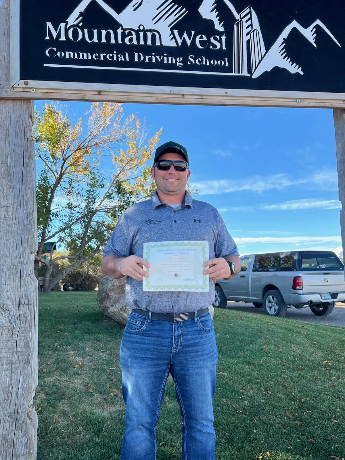 A man wearing sunglasses, a gray polo shirt, and jeans holding a certificate outside near a green lawn with a tree and a vehicle behind him, standing under a sign that reads 'Mountain West Commercial Driving School.'
