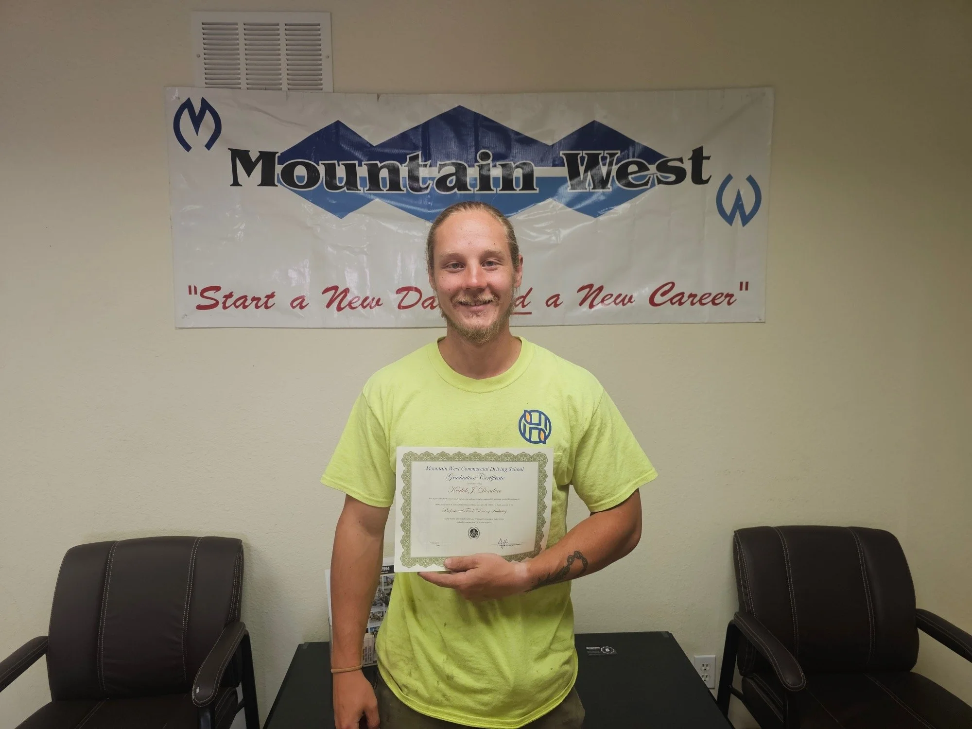 A man standing in front of a Mountain West banner, holding a certificate and smiling. The banner says "Start a New Day and a New Career." The man is wearing a lime green t-shirt with a logo, with two chairs visible beside him.