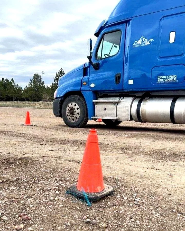 A blue semi-truck parked on a gravel lot with two orange traffic cones, one in front of it and one behind, and trees in the background.