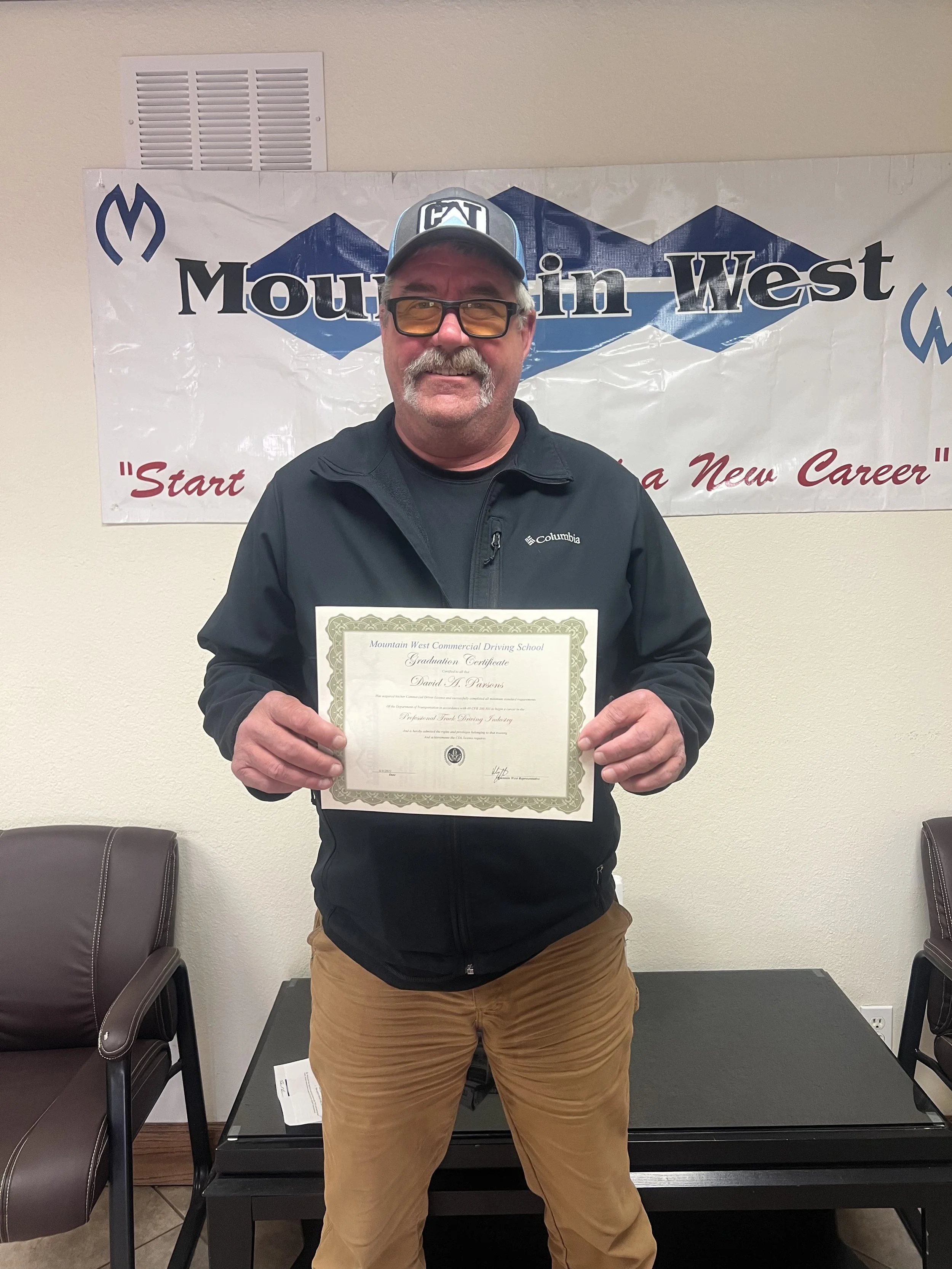 A man standing indoors holding a certificate, wearing a black Columbia jacket, brown pants, a gray baseball cap with a logo, and sunglasses, smiling in front of a Mountain West banner.