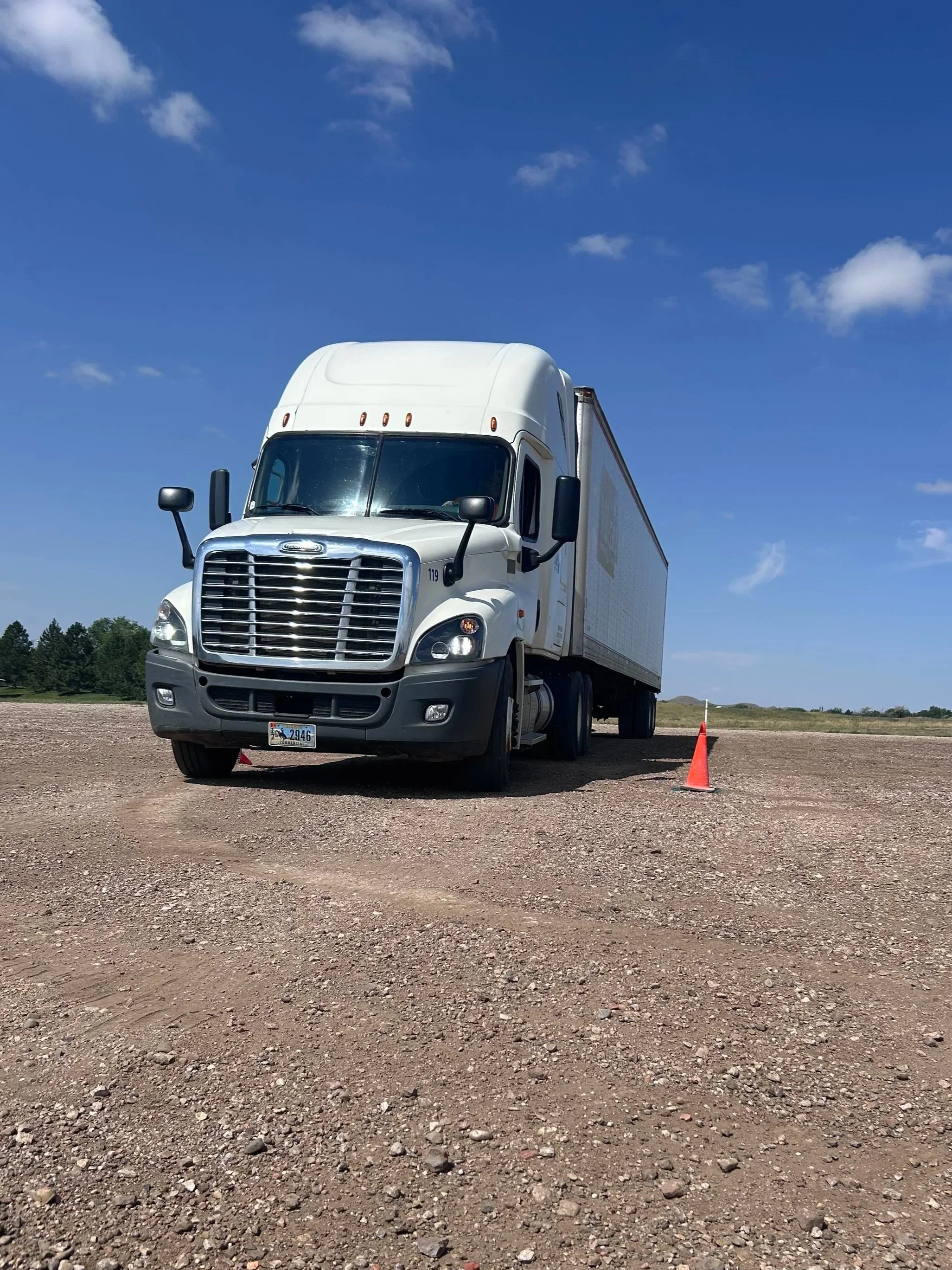 White semi-truck parked on a dirt lot under a blue sky with scattered clouds, with orange traffic cones nearby.