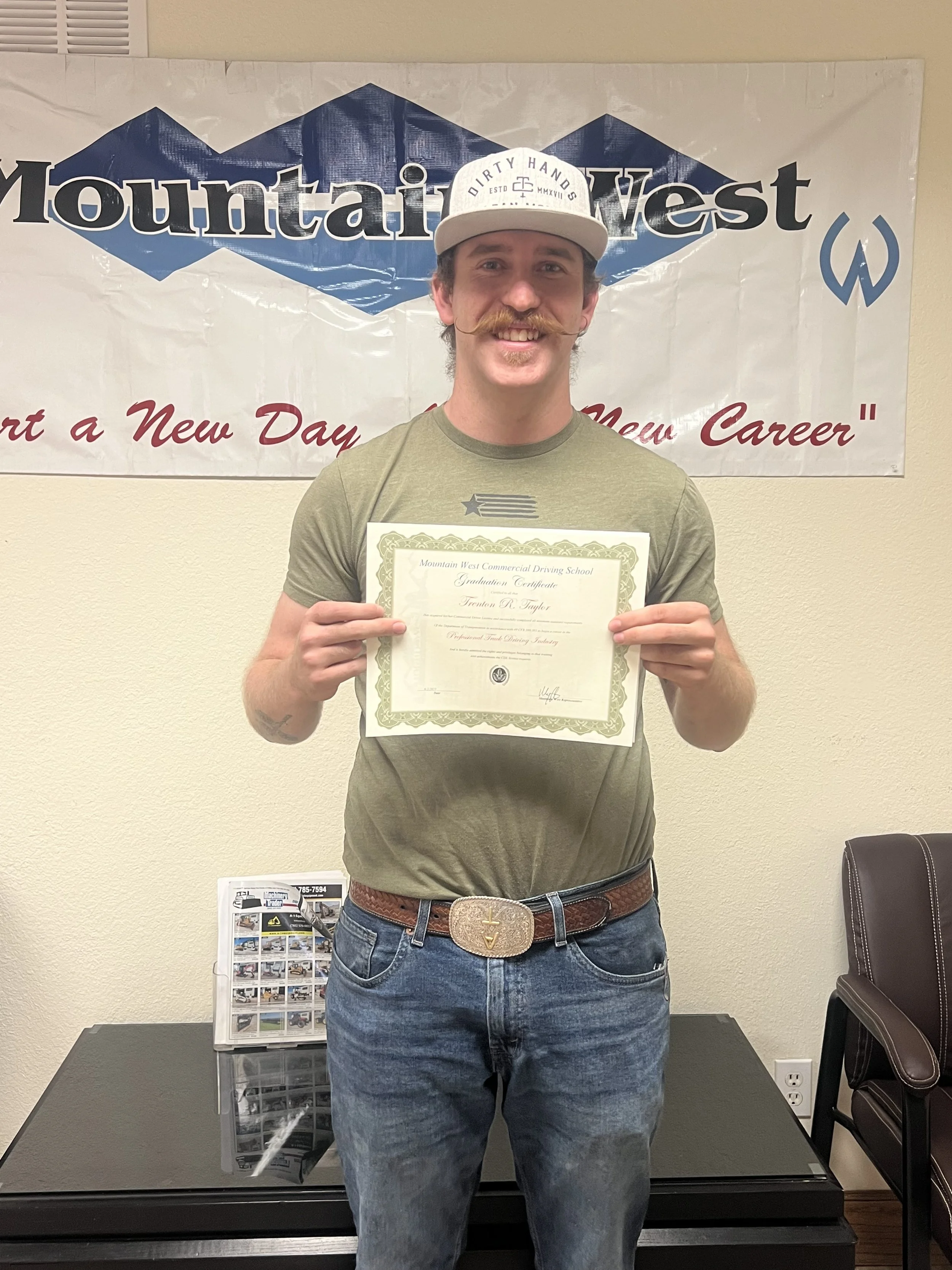 A man standing indoors in front of a Mountain West banner, holding a graduation certificate and smiling.