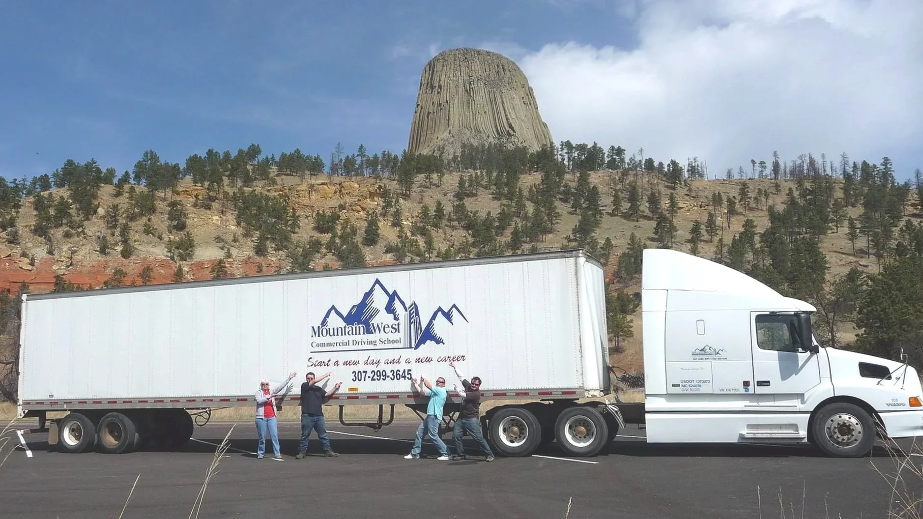 A group of five people standing in front of a large white semi-truck with Mountain West Commercial Driving School logo, in a parking lot with a mountain and a flat-topped rock formation in the background.