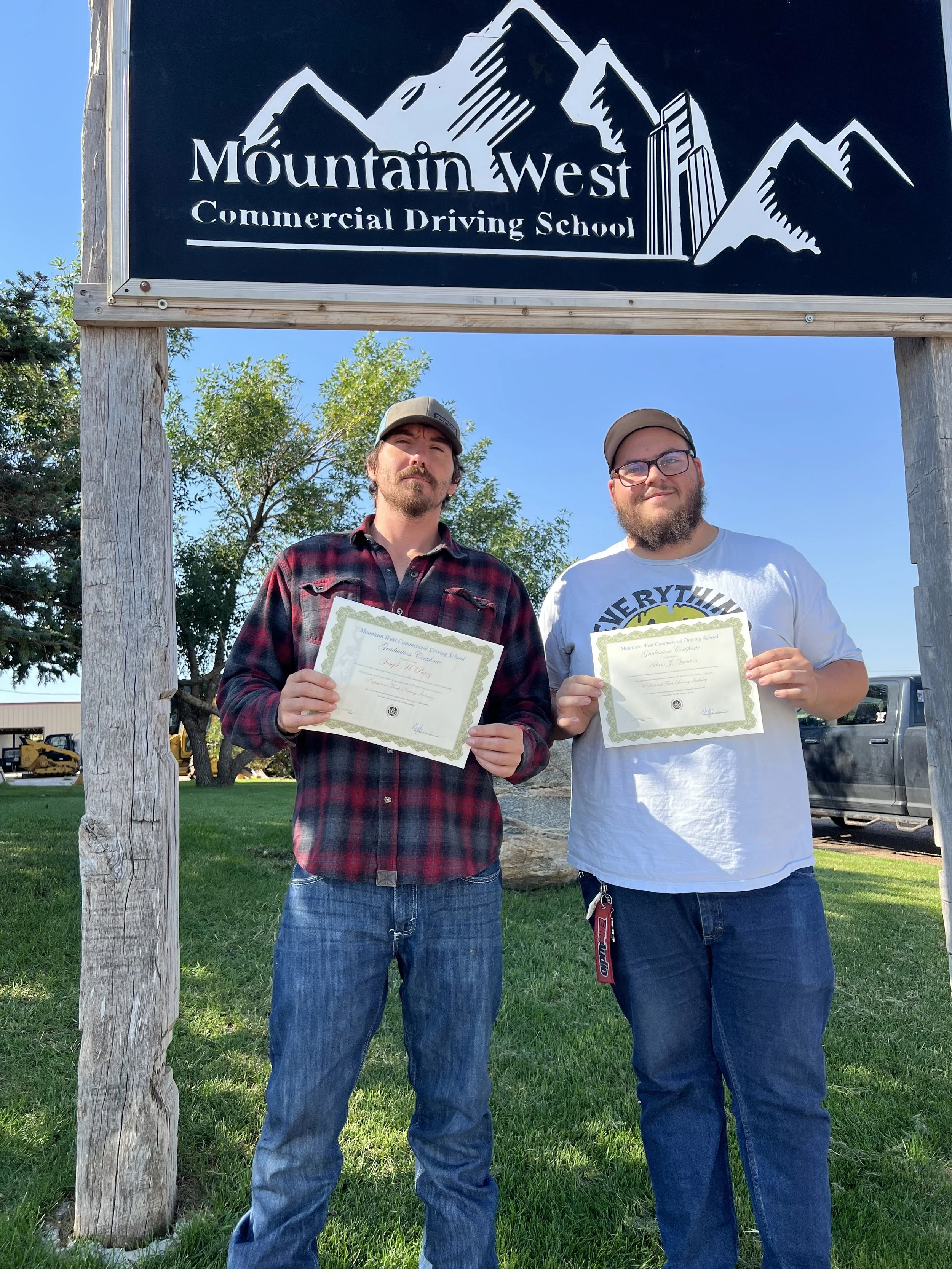 Two men standing outside under a sign that reads 'Mountain West Commercial Driving School,' holding certificates. One is wearing a plaid shirt and the other a white t-shirt.