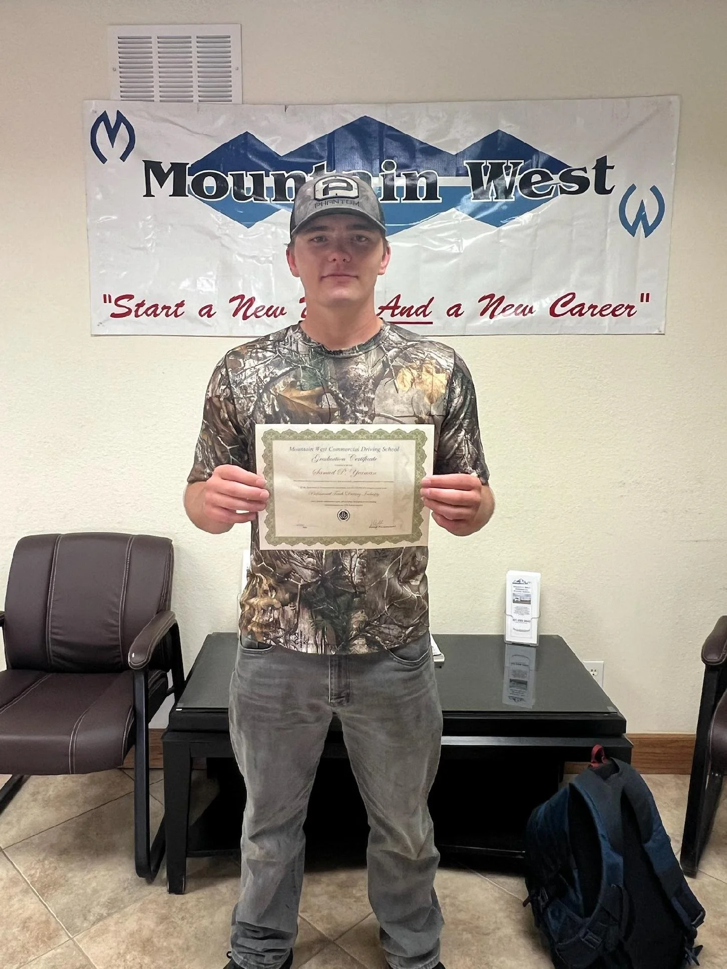 Young man in camouflage shirt and cap holding a certificate in a room with chairs, a table, and a banner that says "Moutain in West" with the tagline "Start a New Job and a New Career."