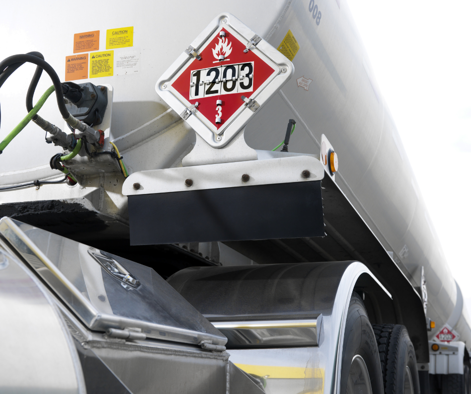 Close-up of a white tanker truck, showing a hazard placard with a flame symbol and number 1203, indicating flammable liquids, along with warning labels and part of the truck's wheel and fender.