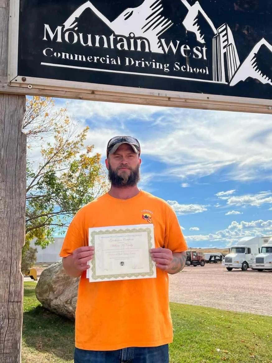 A man with a beard and sunglasses, wearing an orange T-shirt, holding a diploma or certificate, standing outdoors near a wooden post with a mountain-themed sign for Mountain West Commercial Driving School. There are vehicles and a cloudy sky in the b