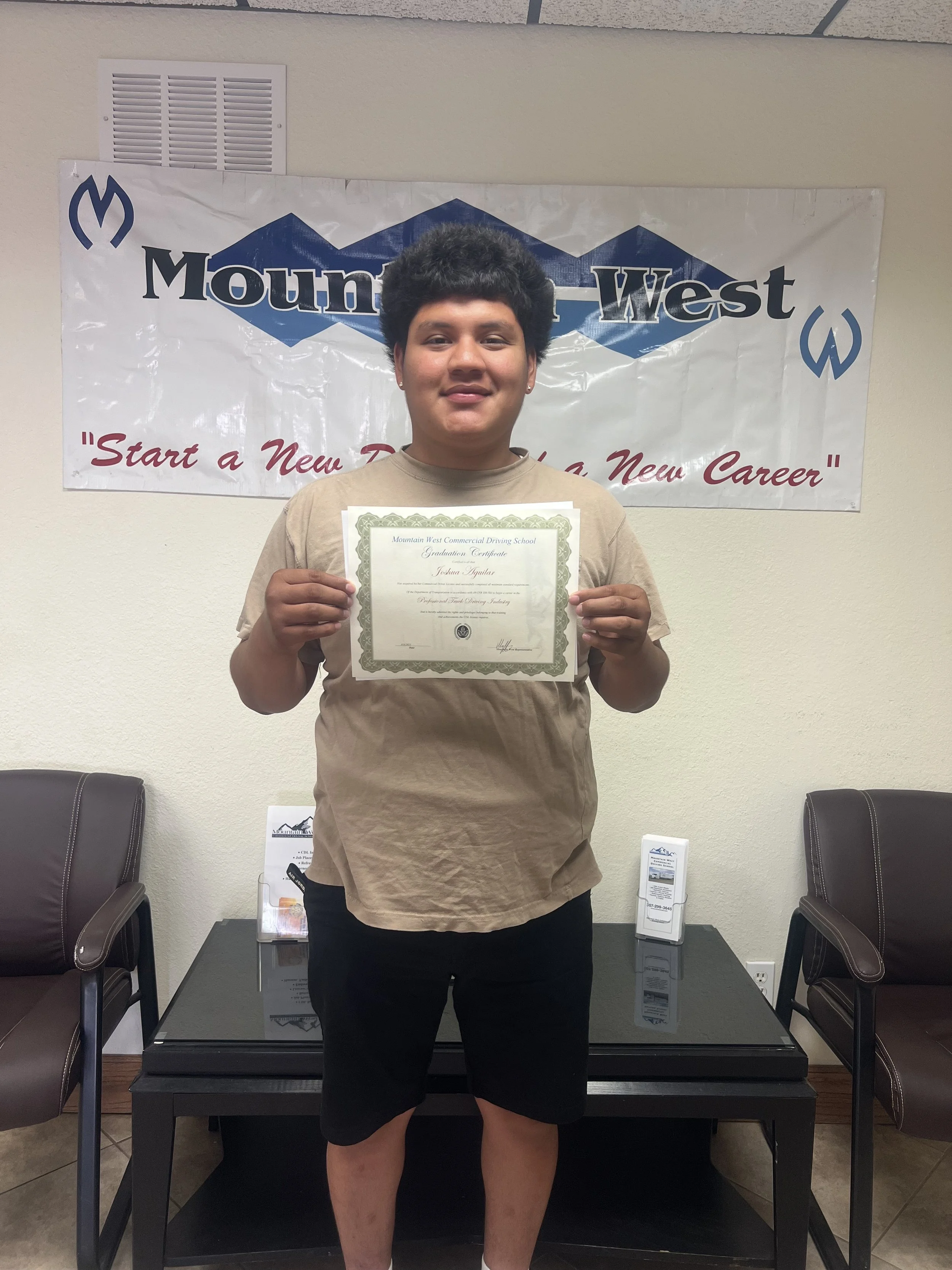 Young male holding a graduation certificate in front of a Mountain West banner, standing between two chairs in an office setting.