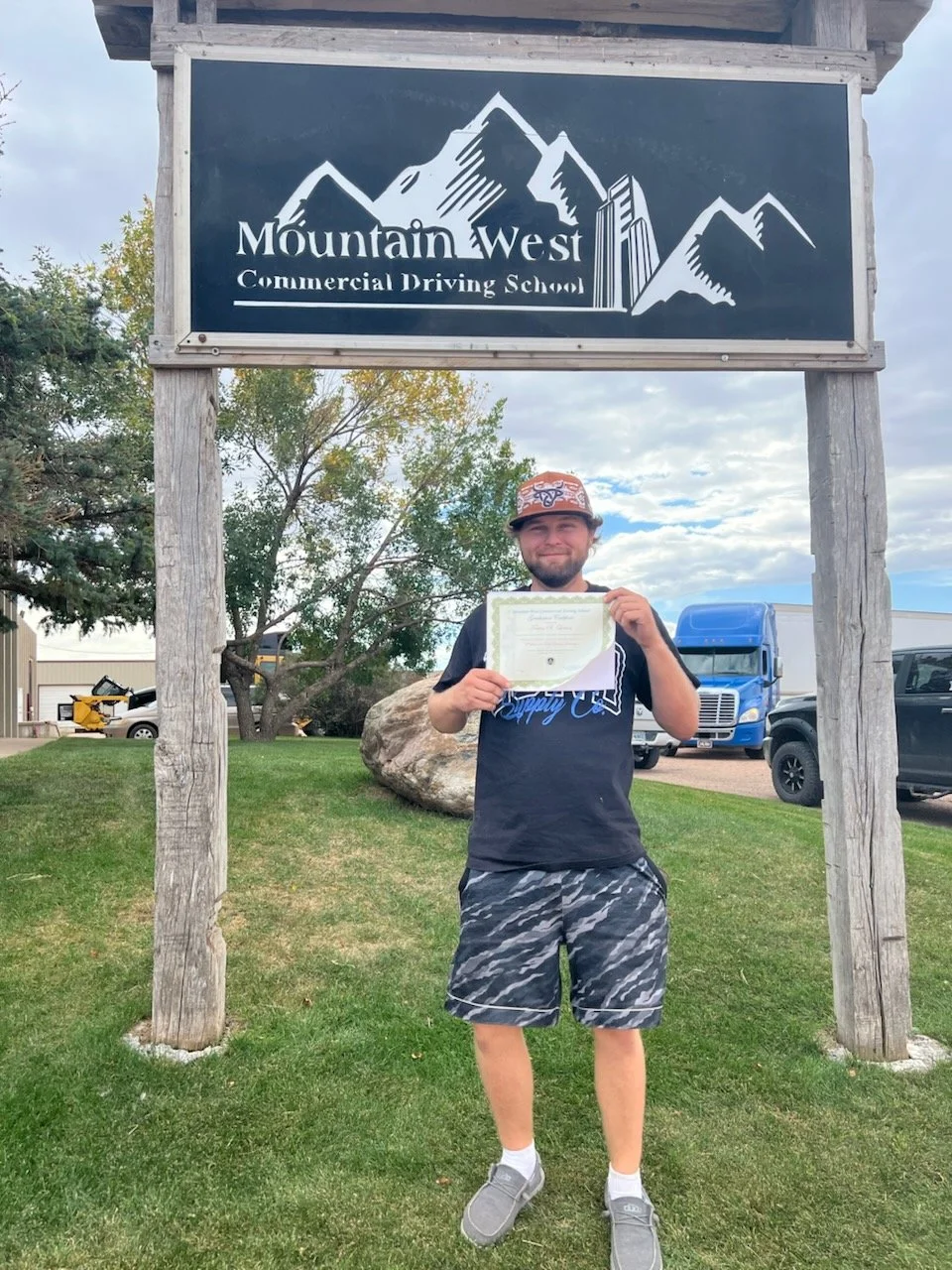 A young man standing on grass holding a certificate in front of a mountain-themed sign that reads 'Mountain West Commercial Driving School.' He is smiling, wearing a patterned cap, a black t-shirt, camouflage shorts, and gray shoes. There are trucks 