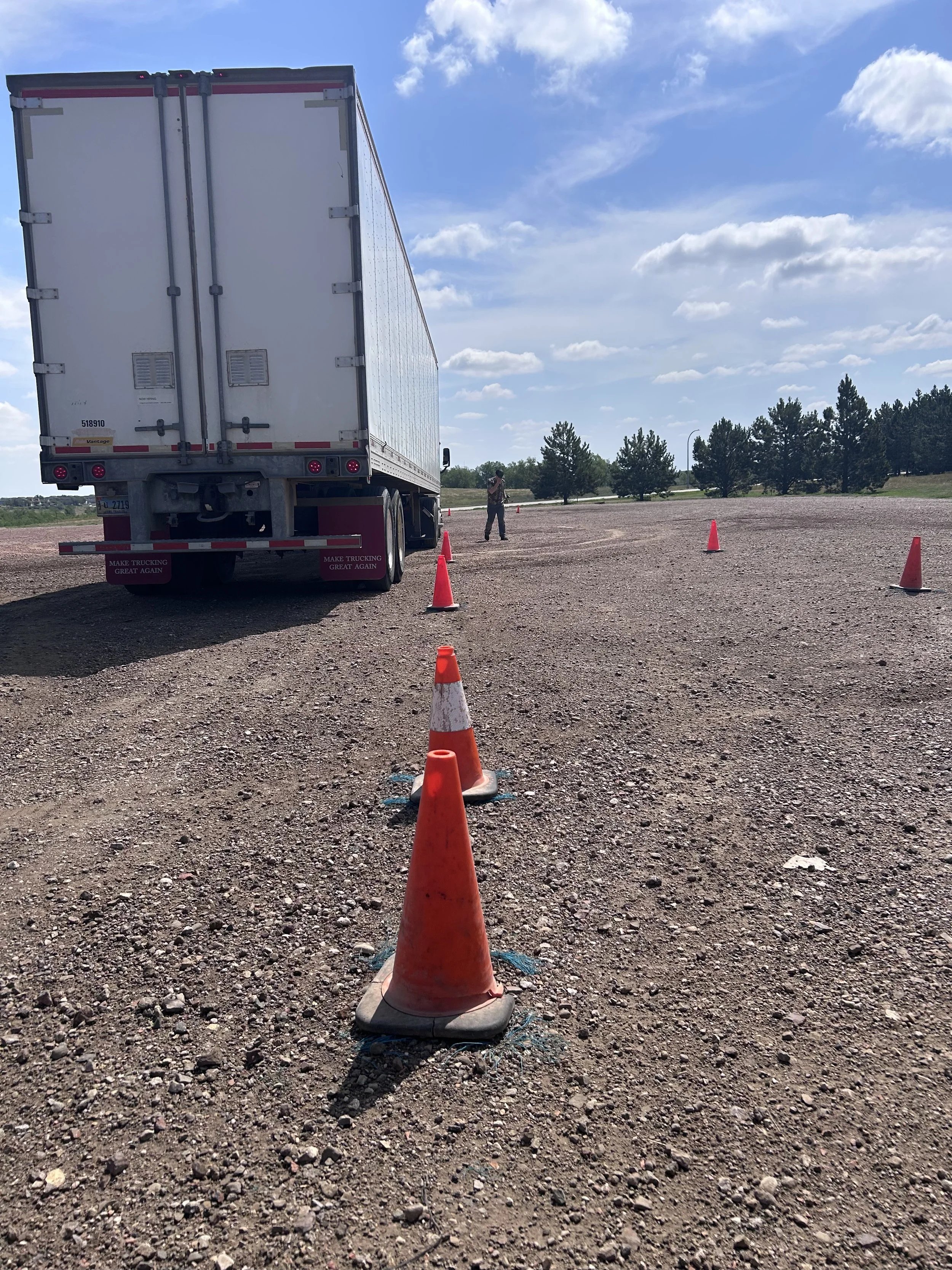 A large semi-truck parked on a gravel lot with orange traffic cones arranged around it, and a person taking a photo in the background with a blue sky and green trees.