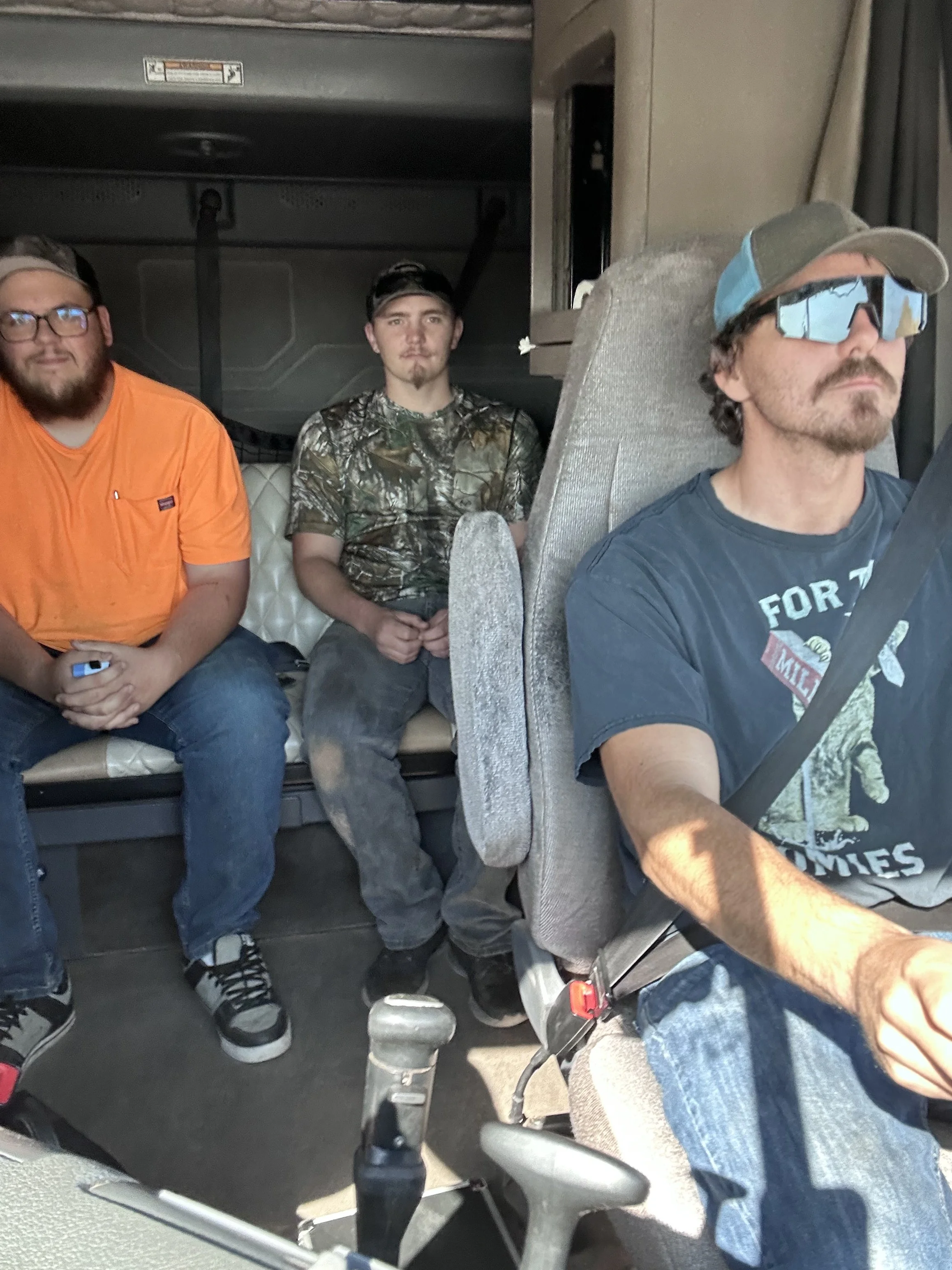 Three men sitting inside a vehicle, with one man in the driver's seat wearing sunglasses and a cap, and two men sitting behind him—one in an orange shirt and the other in a camouflage shirt.