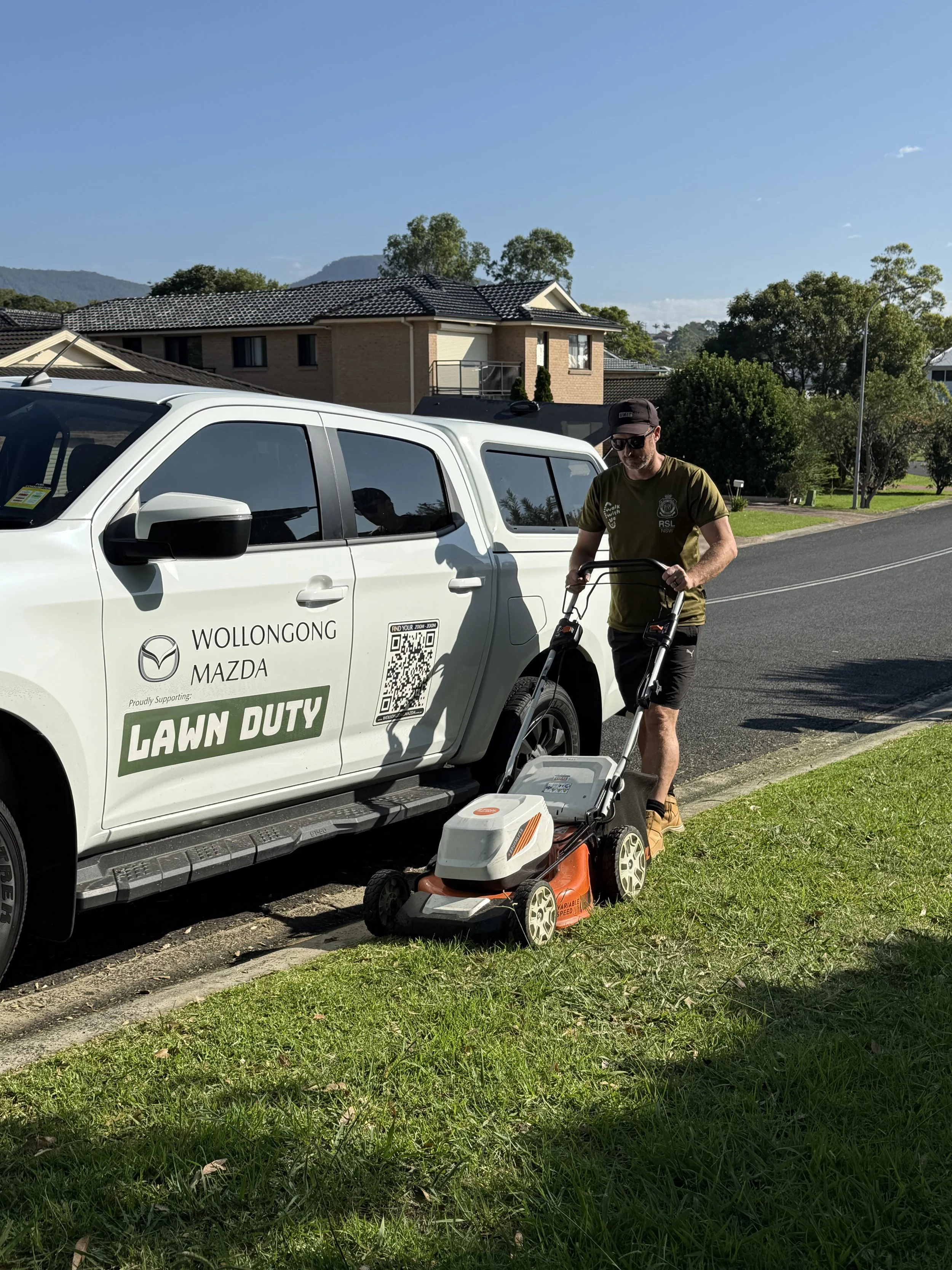 A man mowing the lawn next to a white Mazda vehicle parked on the street in a residential neighborhood with houses and trees in the background.