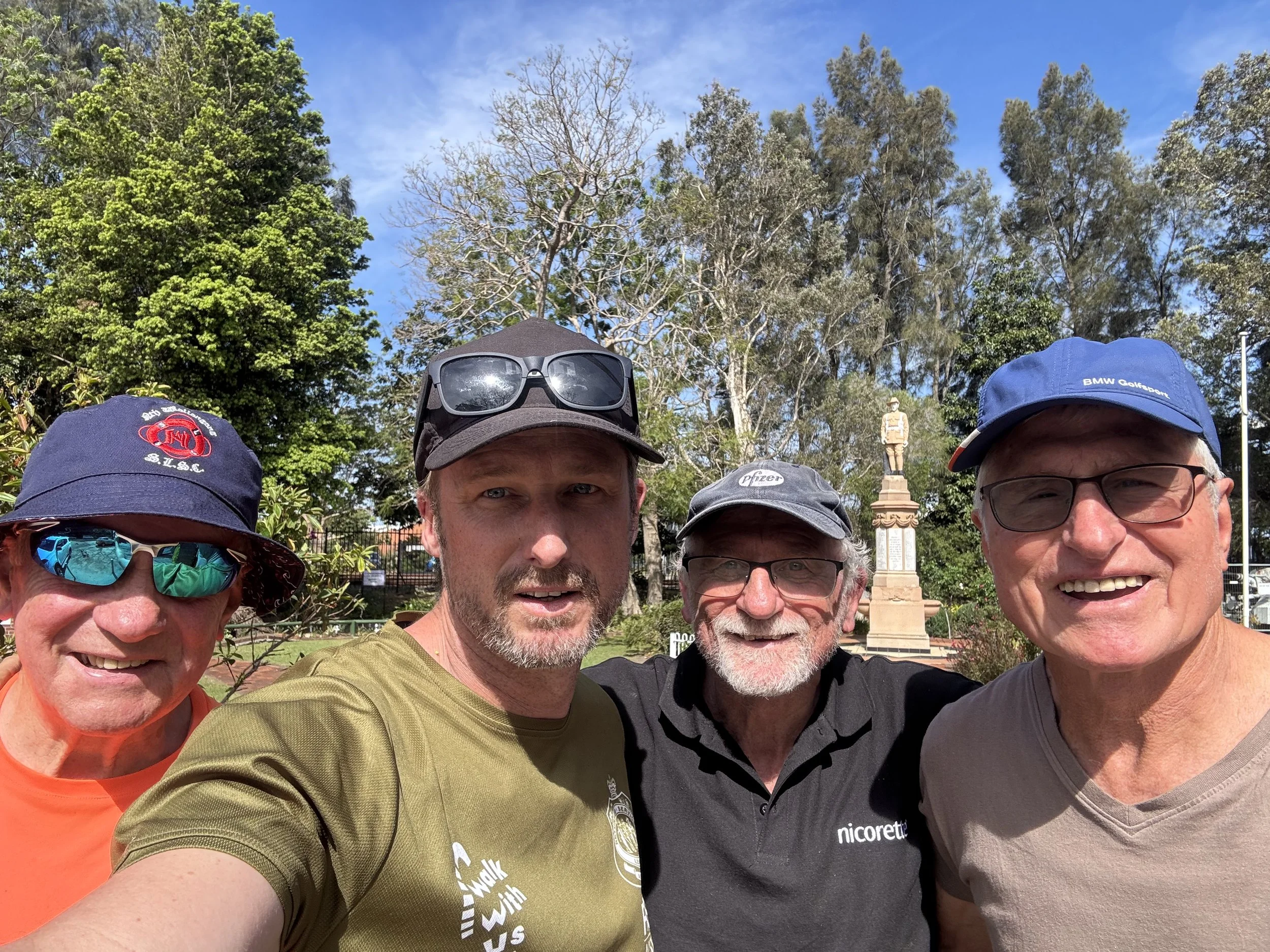 Group of five people taking a selfie outdoors with trees and a monument in the background.