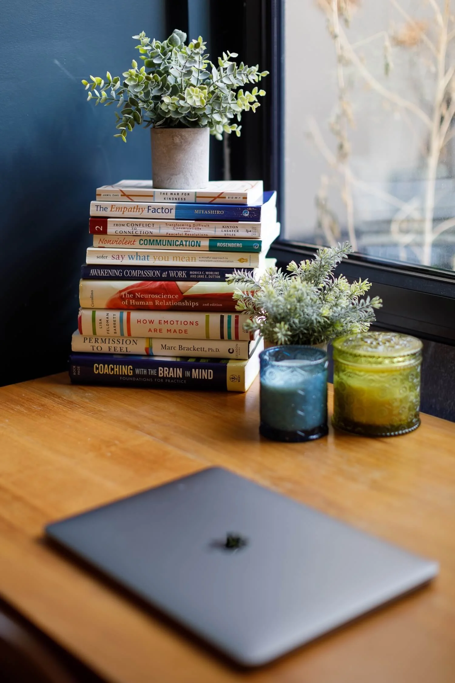 An laptop on a tidy desk with a stack of books relating to topics such as empathy, communication and emotions.