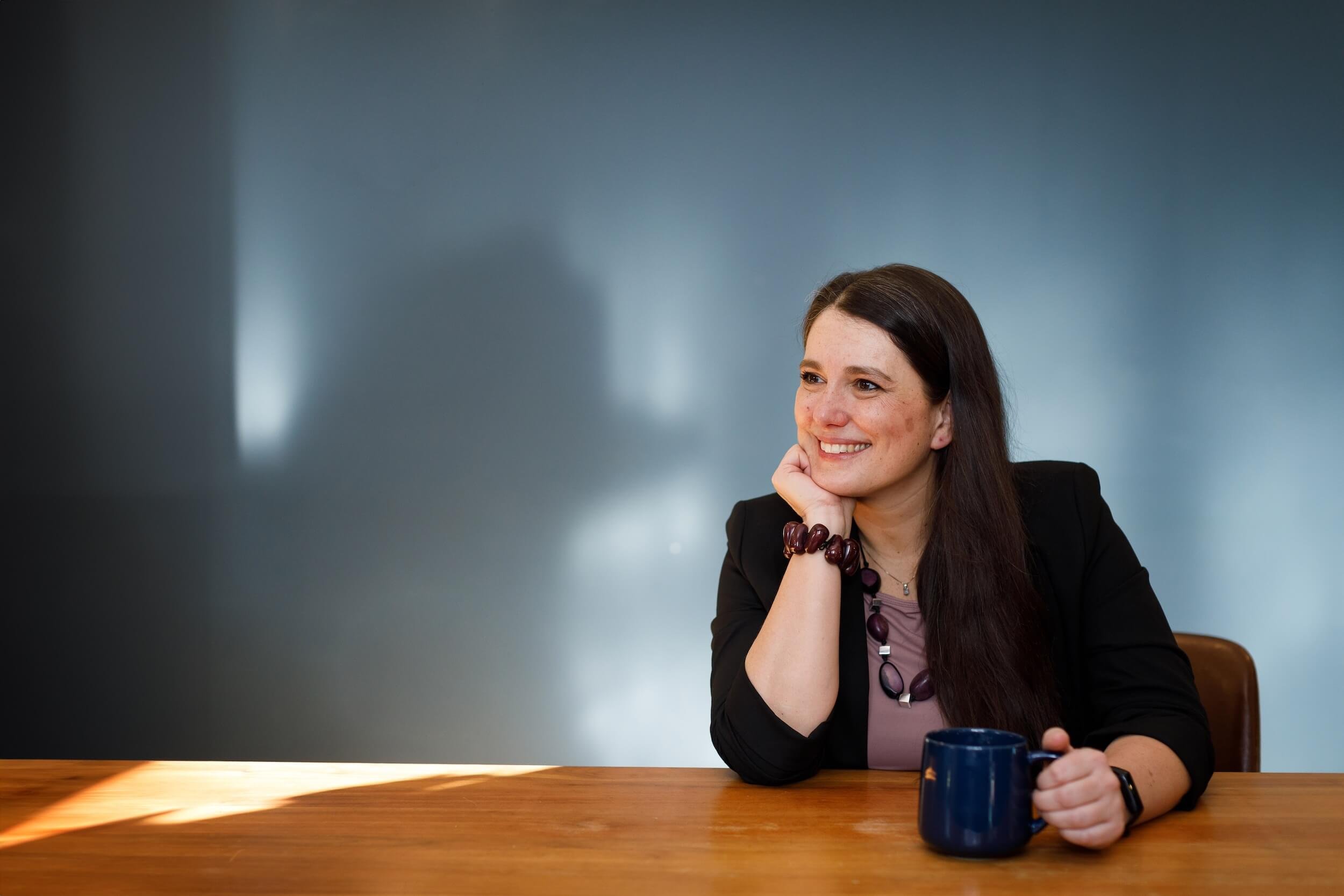 Clara Moisello seated at a wooden table, smiling warmly with a ceramic mug — inviting open, compassionate conversation