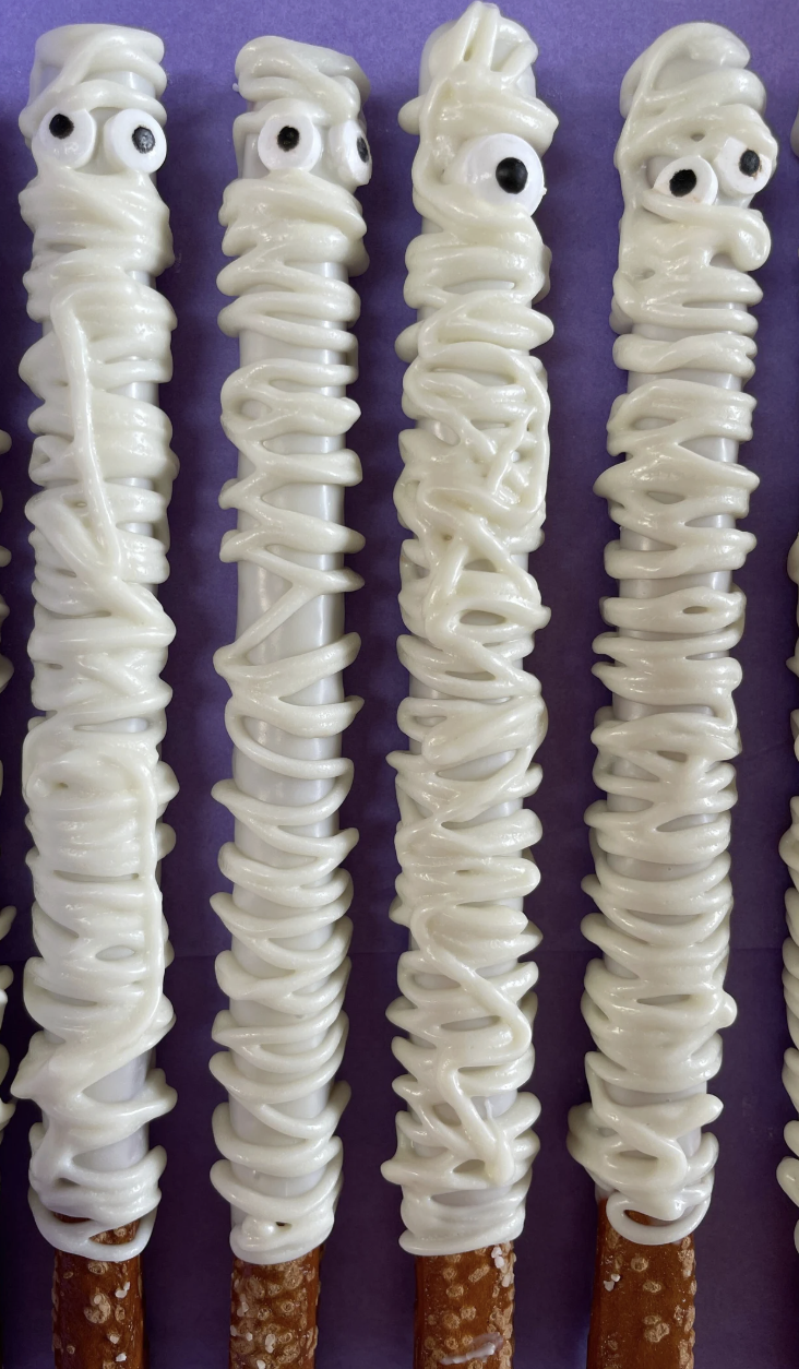 Four pretzel sticks dipped in white chocolate, decorated with black and white candy eyes to resemble mummies, placed against a purple background.