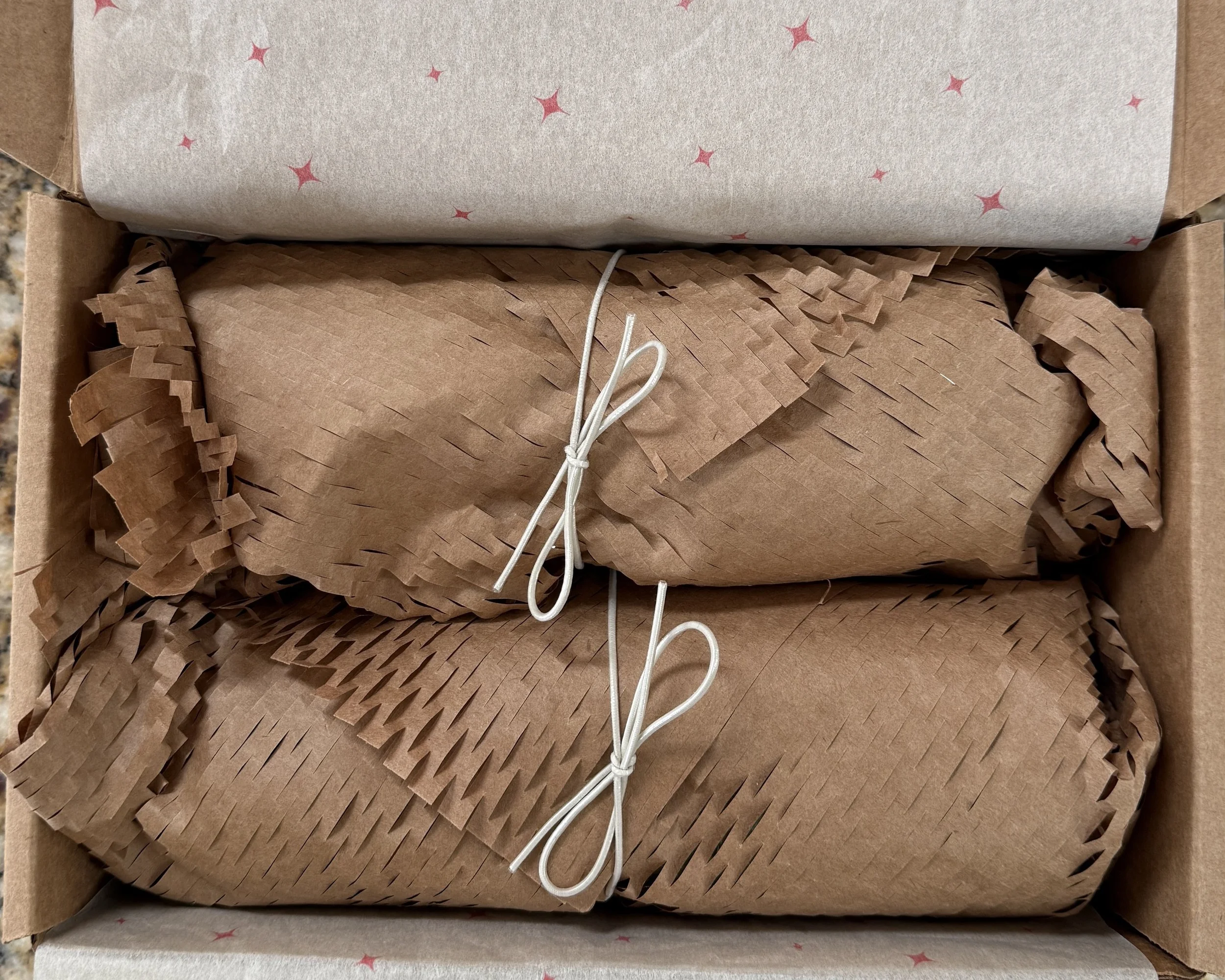 The image shows packaged chocolate-dipped pretzels wrapped in recycled brown packing paper, white string tied around the paper, and a piece of branded tissue paper with small pink star patterns, partially visible at the top.