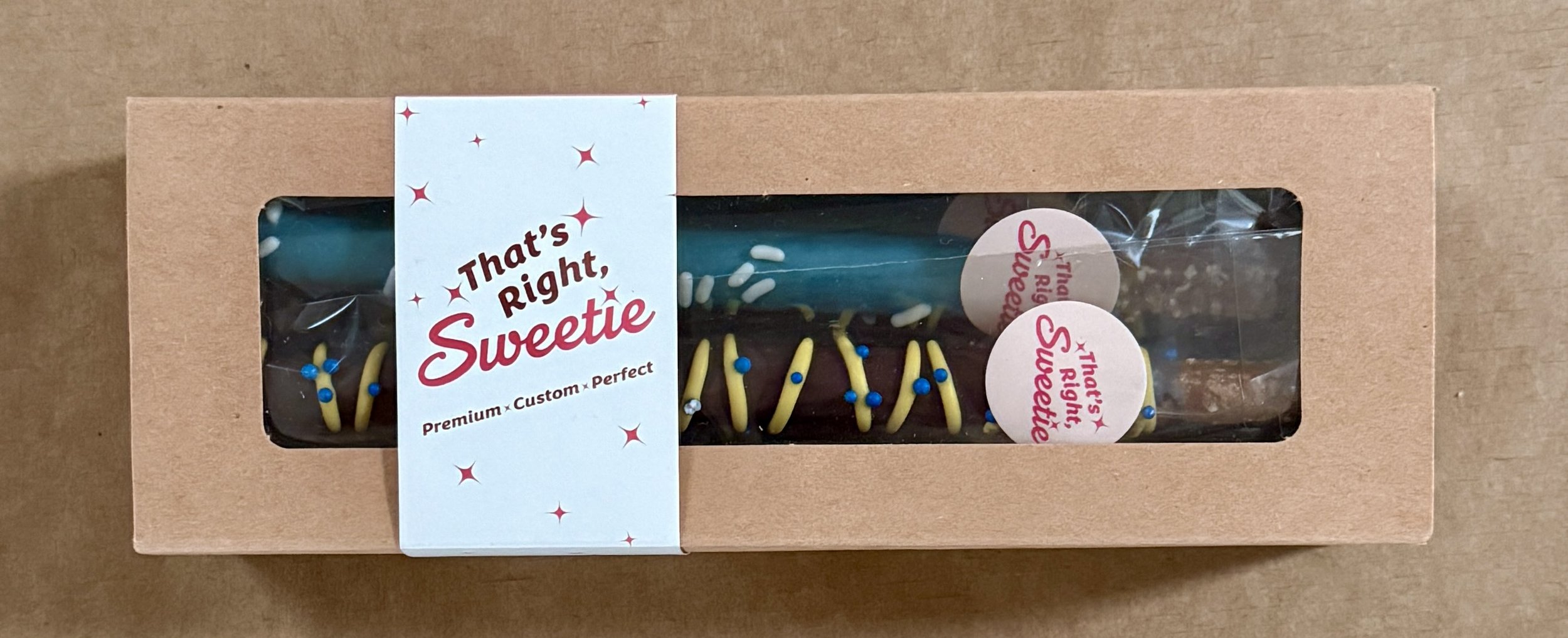 A rectangular birthday cake with blue and yellow icing decorations and a matchstick candle inside a clear plastic window in a cardboard box. The box has a label that reads 'That's Right, Sweetie' and 'Premium Custom Perfect'.