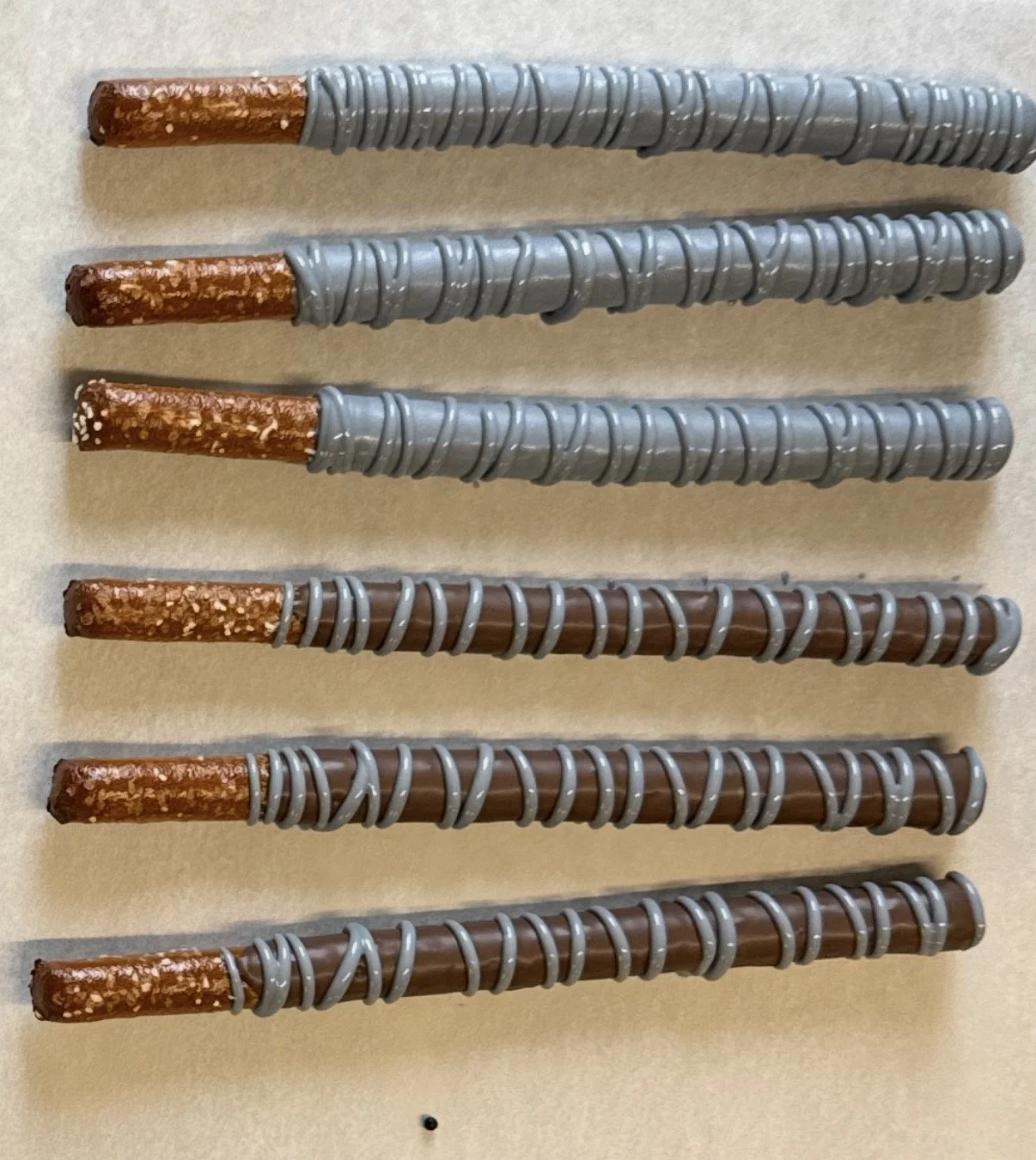 Six pretzel rods dipped in chocolate and decorated with branded, corporate colors, gray drizzle, arranged parallel on a recycled packaging surface.