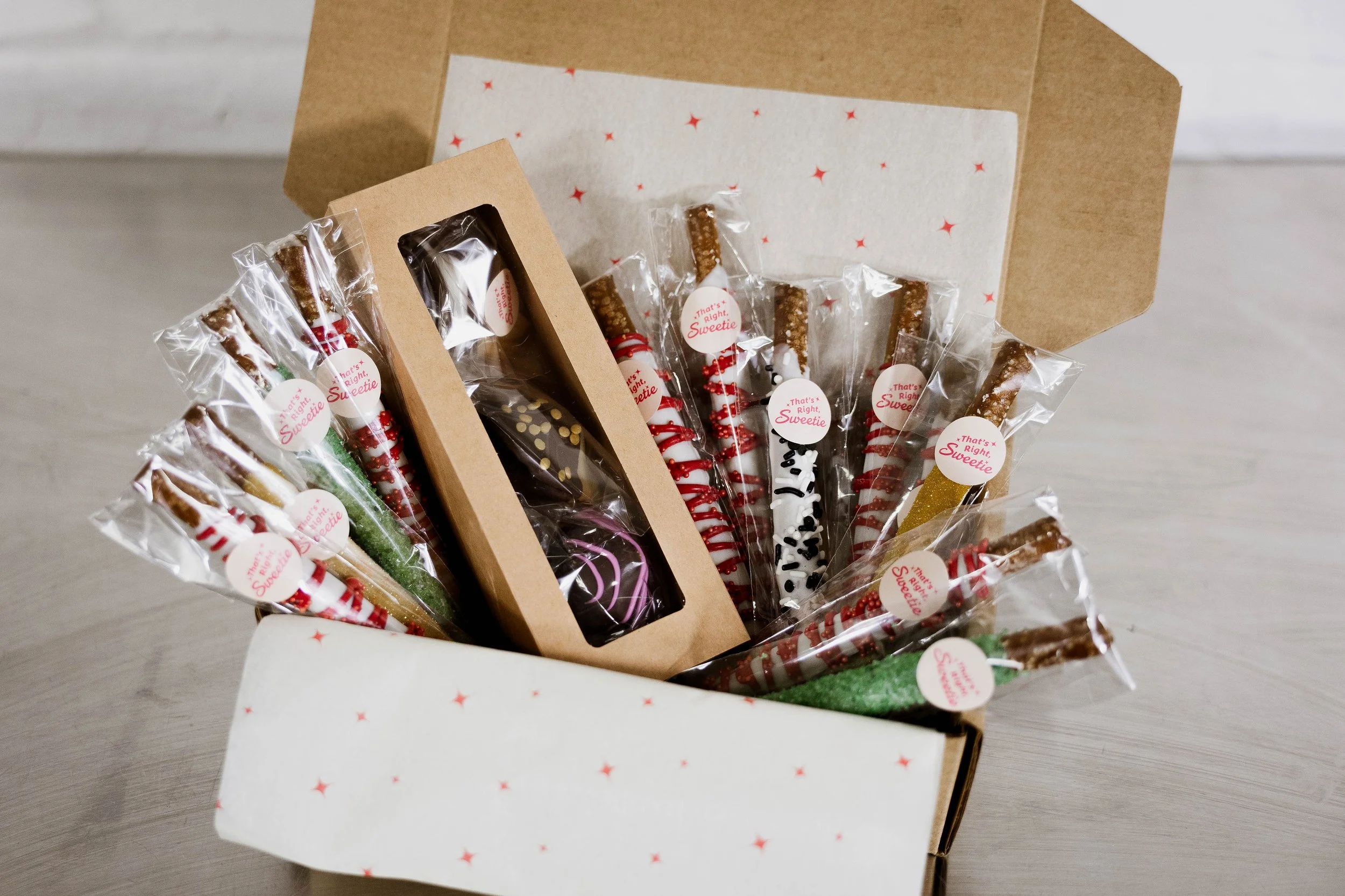A decorated box filled with assorted holiday treats including chocolate-covered pretzel rods wrapped in clear plastic with festive stickers, on a light-colored surface.