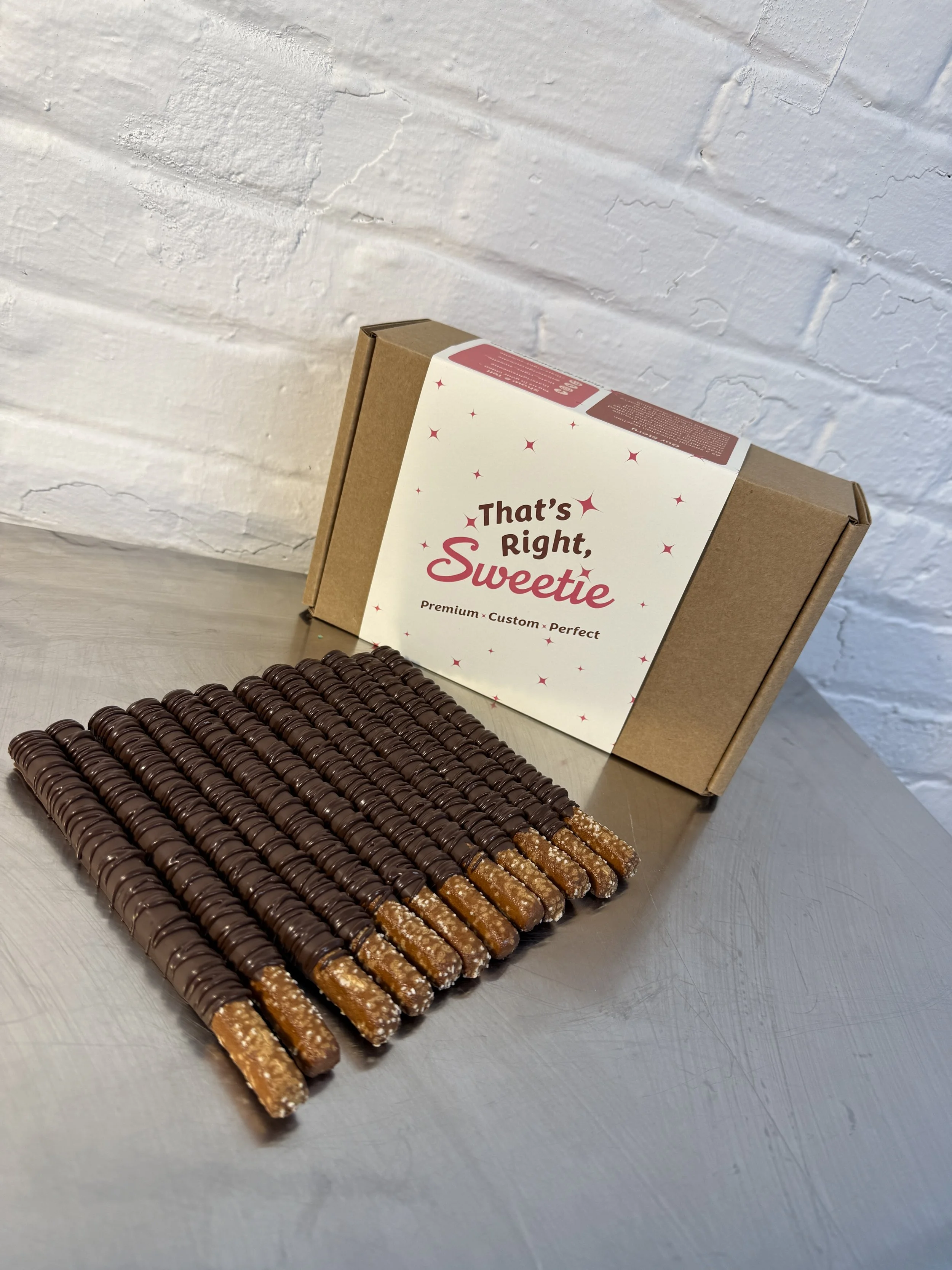 Rows of chocolate-covered wafer sticks arranged on a silver tray, with a cardboard box labeled 'That's Right, Sweetie' in the background, against a white textured brick wall.