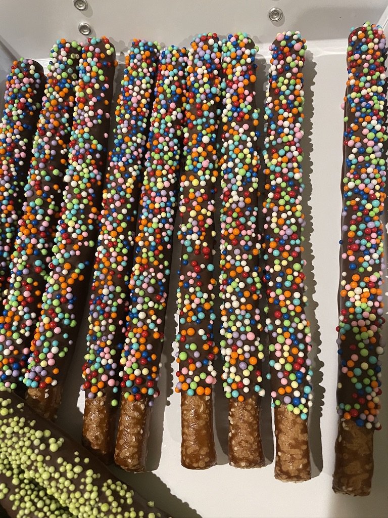 Chocolate pretzel rods covered in colorful round sprinkles.