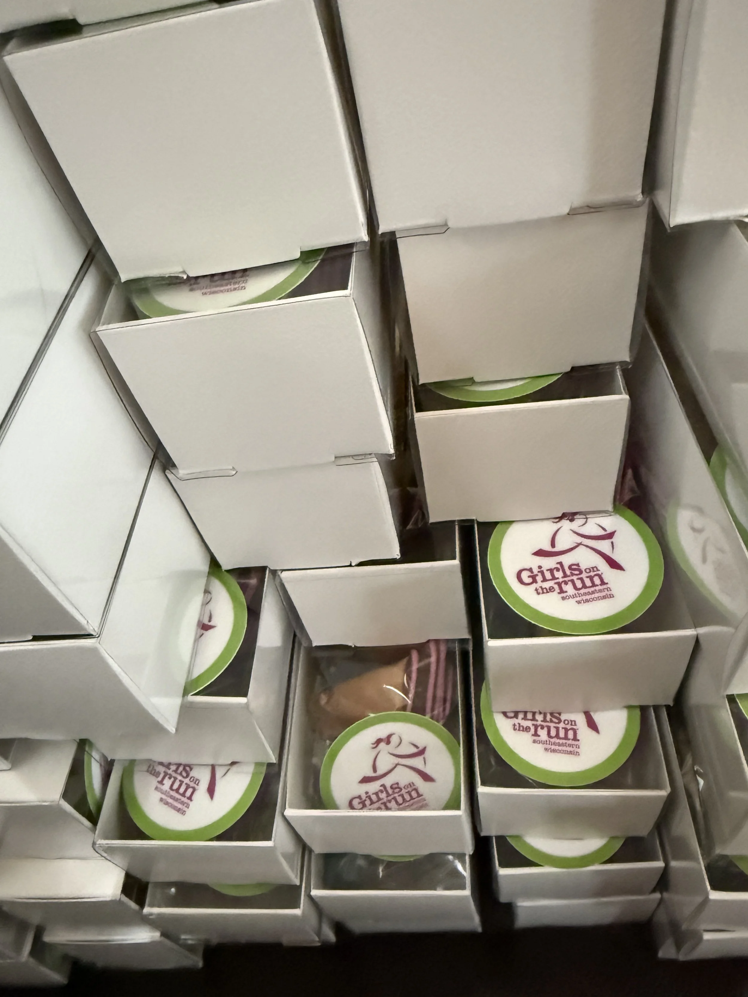 Multiple stacked white boxes with circular stickers inside. The stickers have a woman running and the text 'Girls on the Run' in a purple font with a green border.