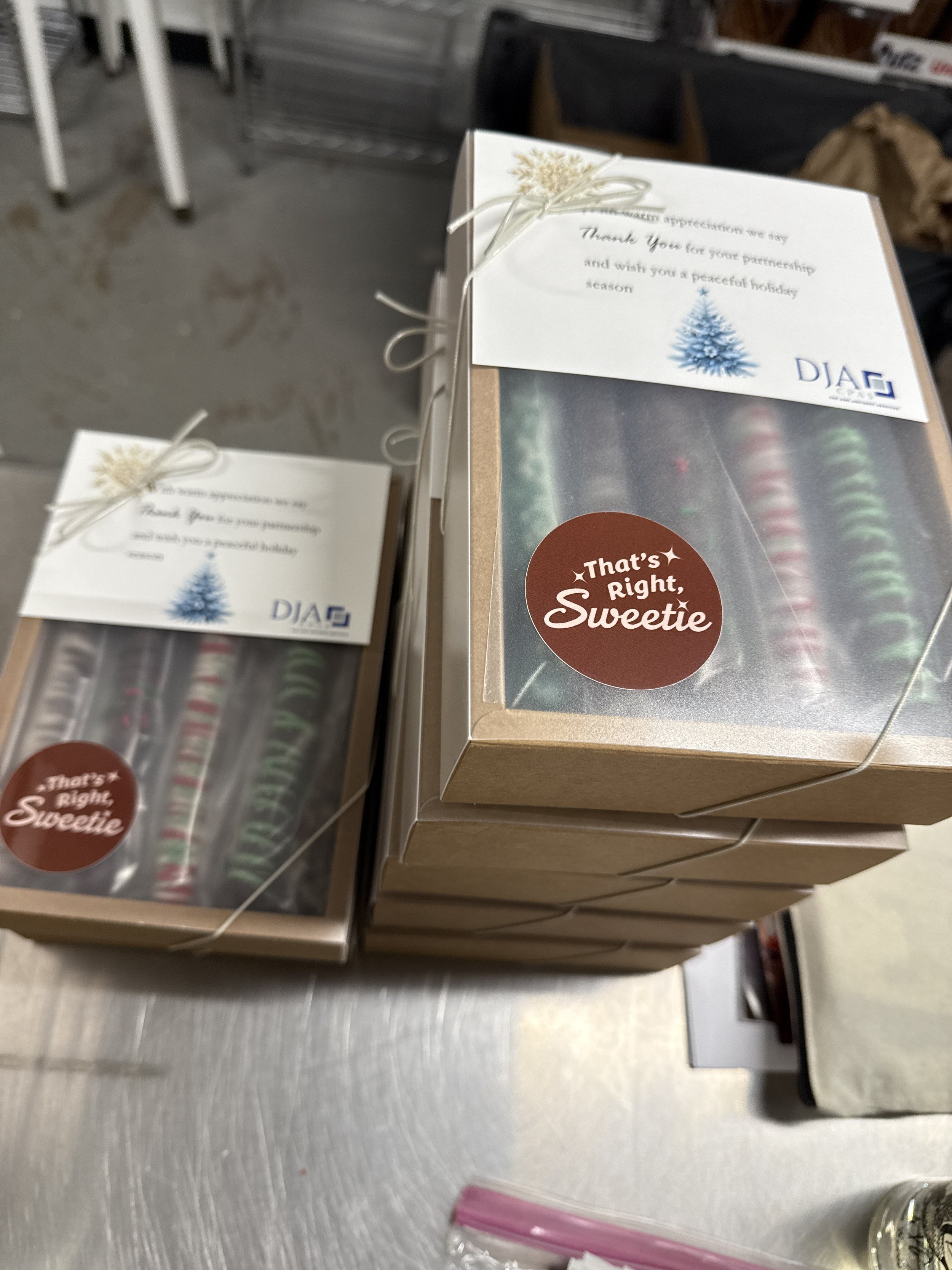Close-up of chocolate-dipped holiday-decorated pretzel rods packaged to send as corporate gifts. Tied with a string and a sticker that says "That's Right, Sweetie." The individually packaged one-dozen gifts are stacked on a metal surface in a store.