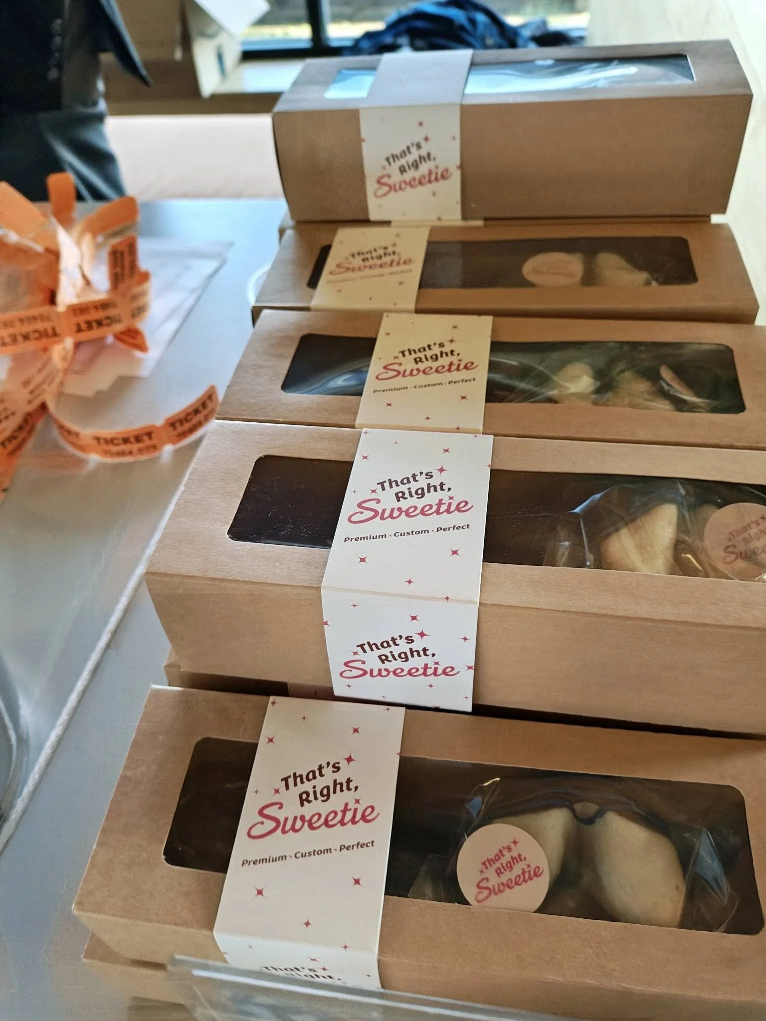 Three wooden boxes with transparent glass windows containing cookies, stacked on a table. Each box has a white paper band with red and black text that reads 'That's Right, Sweetie,' and additional small text. To the left, there is a partial view of o