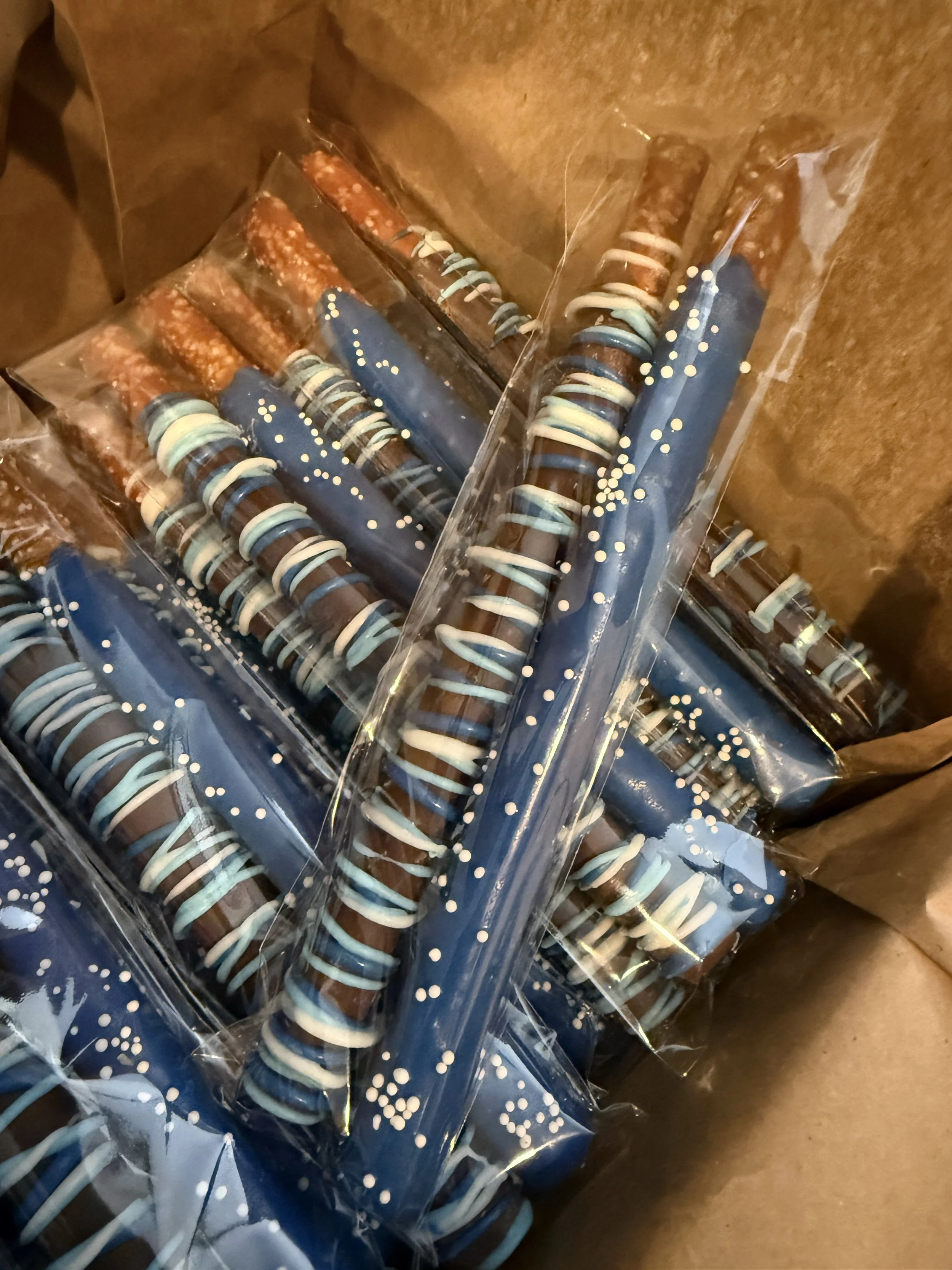 Close-up of wrapped candy or chocolate sticks in blue and brown colors with white and light blue ribbon-like decorations, inside clear plastic packaging.