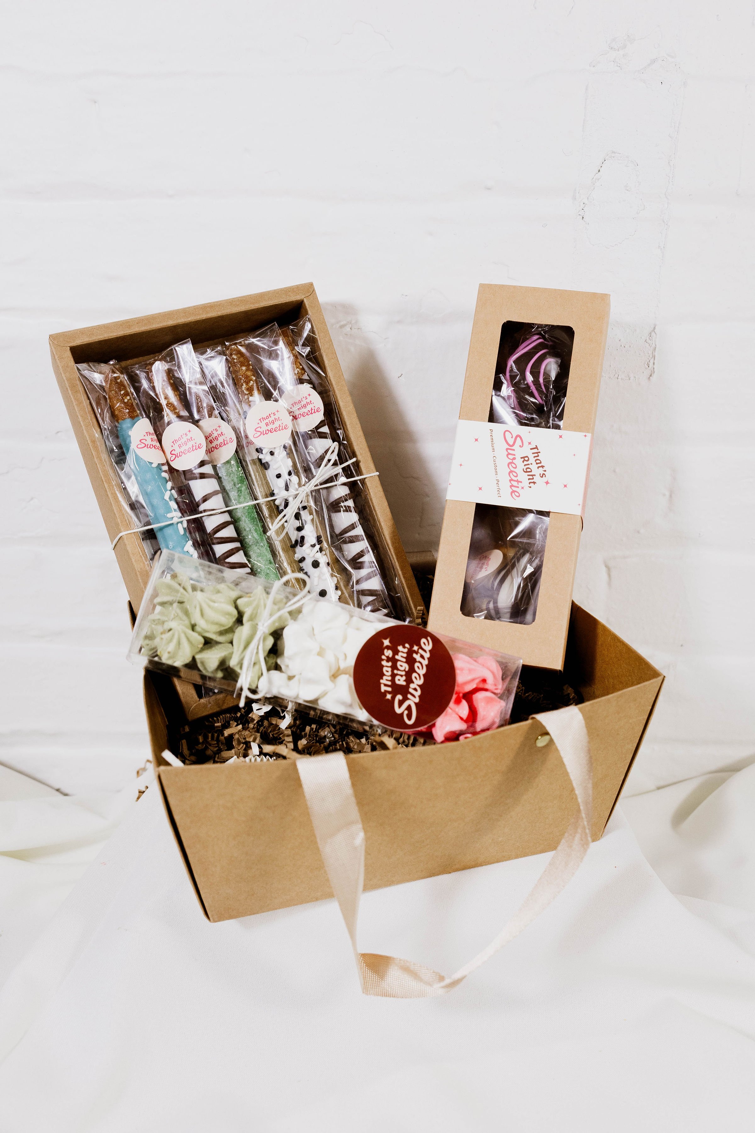 A brown gift box with a white ribbon contains assorted candy and treats, including decorated cookies and marshmallows, set on a white surface against a white brick wall background.