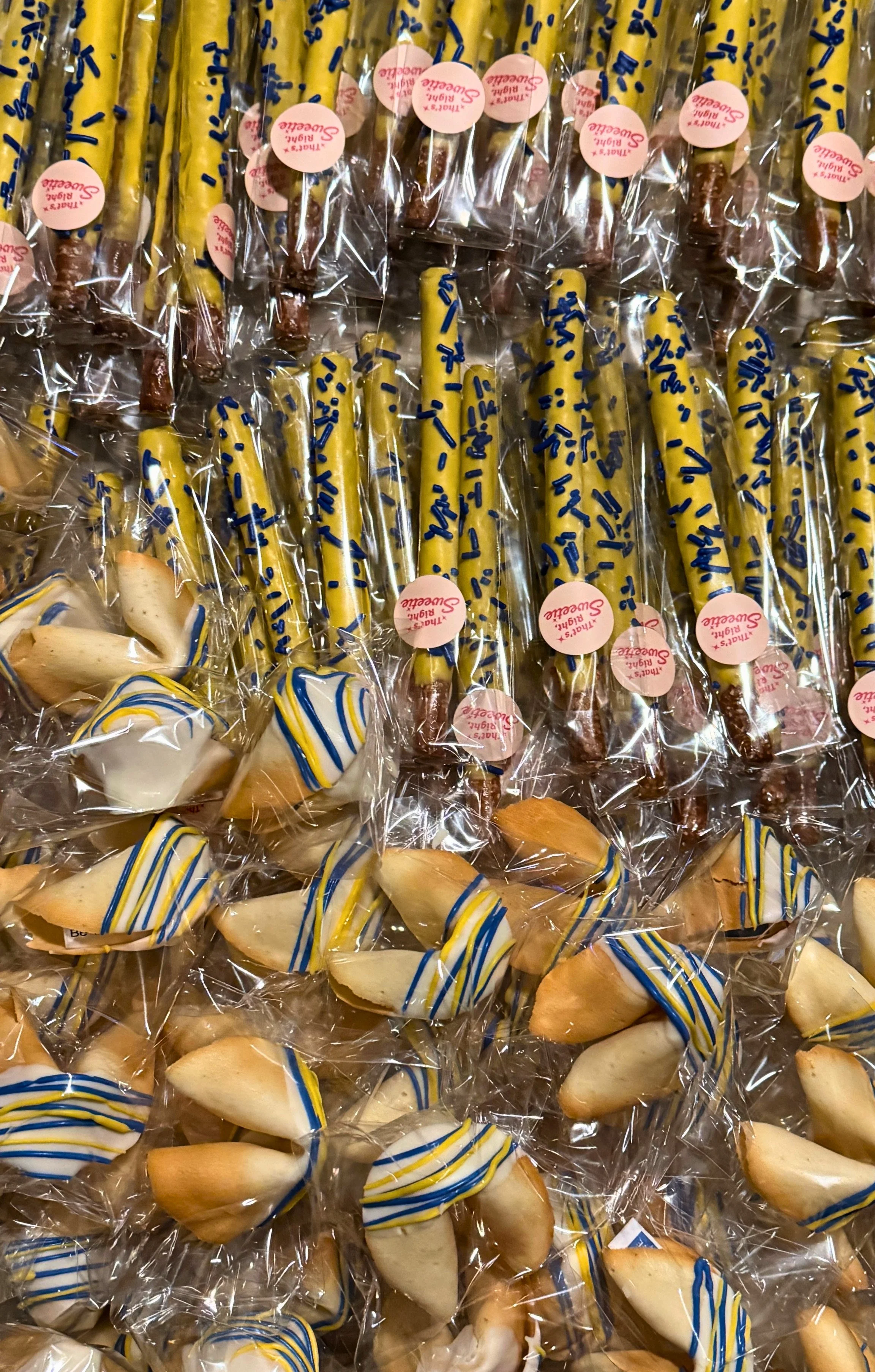 Assorted cookies and candies wrapped in clear plastic, decorated with blue, yellow, and white icing, some with chocolate and pink labels.