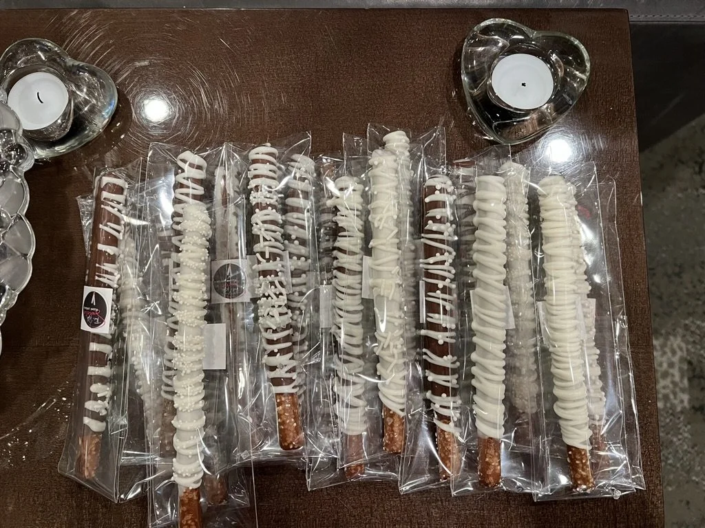 Nine chocolate-covered pretzel sticks with white icing drizzle, individually wrapped in clear plastic, arranged on a wooden surface with two heart-shaped candles nearby.