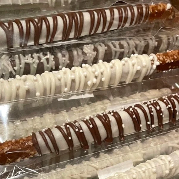 Close-up of chocolate-covered pretzel rods decorated with white and dark chocolate drizzles and sprinkles, stored in clear plastic packaging.