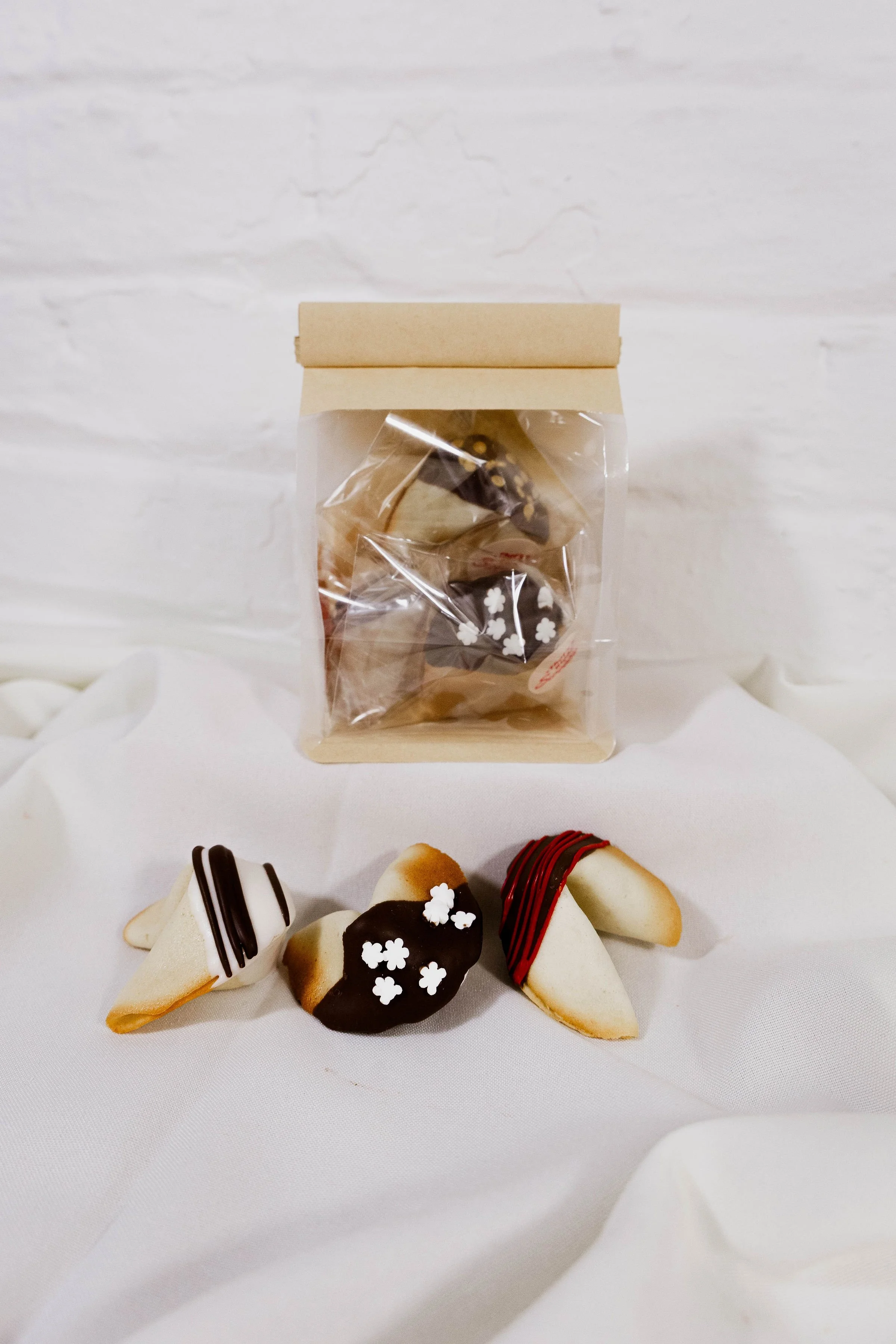 Assorted cookies with various chocolate and white icing decorations, some with flower-shaped sprinkles, arranged on a white surface with a small bag of cookies in the background.