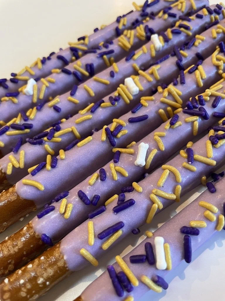 Close-up of three pretzel sticks coated in purple chocolate and sprinkled with purple, yellow, and white sprinkles.