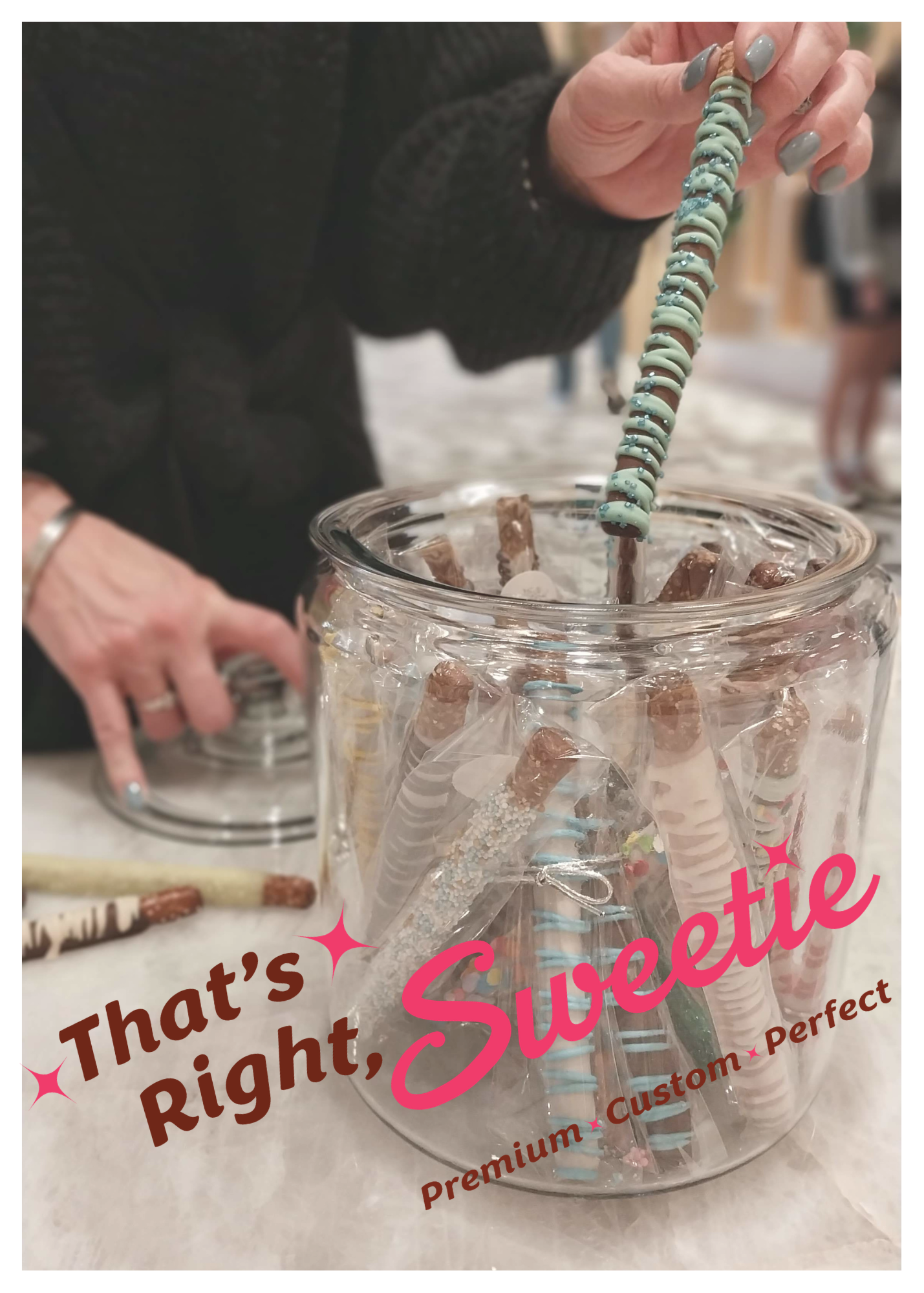 A person with painted nails holds a decorative item over a glass jar filled with wrapped chocolate sticks, with more chocolate sticks and a sign with partially visible words on the table.
