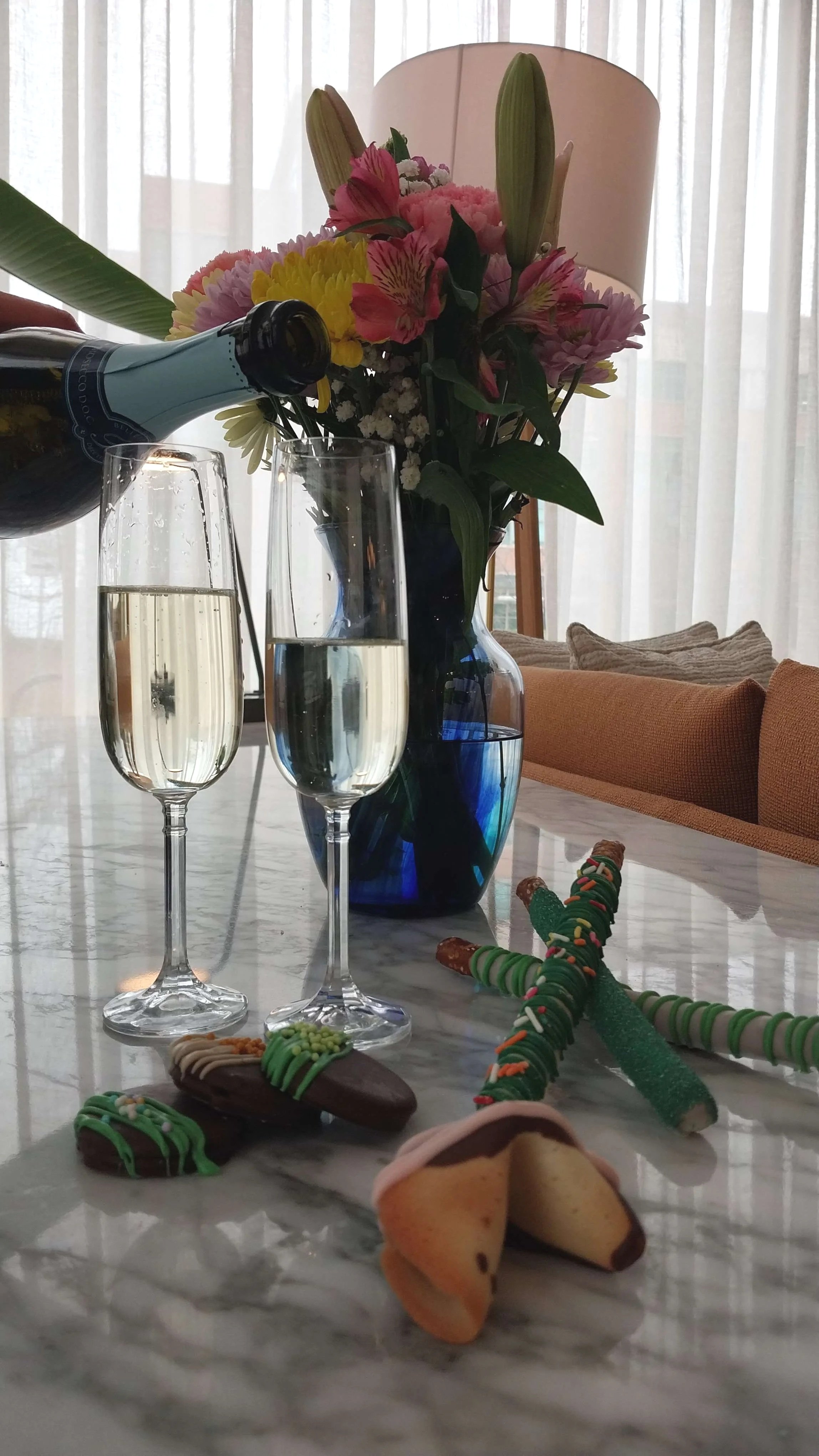 A table with two glasses of champagne, a blue vase with pink, yellow, and white flowers, a bottle pouring champagne, and assorted decorated Christmas cookies. In the background, a lamp and curtains are visible.
