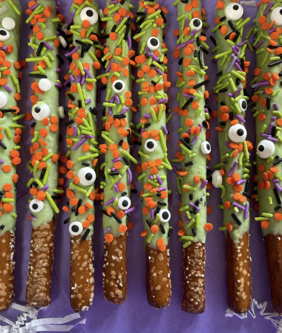 Pretzel sticks dipped in green icing and decorated with orange, purple, black, and white sprinkles, some resembling eyes, arranged on a purple surface.