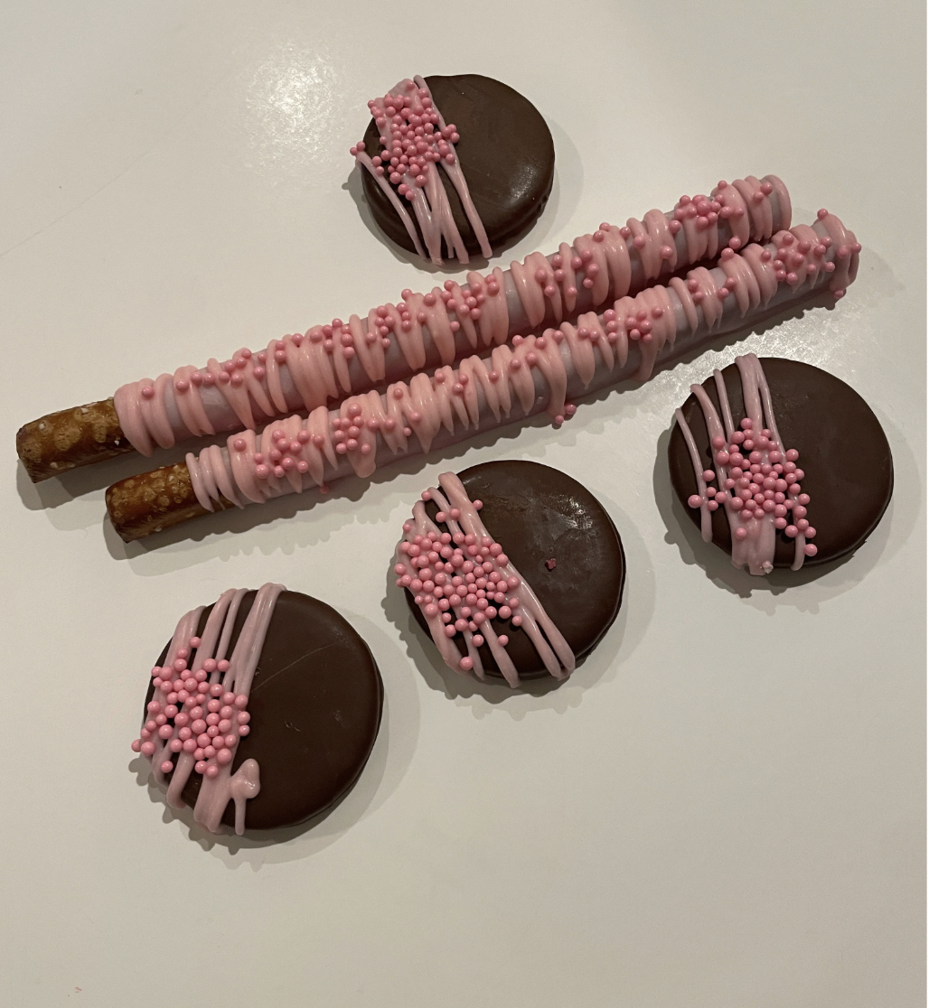 Two pink and brown decorated pretzel sticks and four chocolate-covered cookies with pink and brown sprinkles on a white surface.