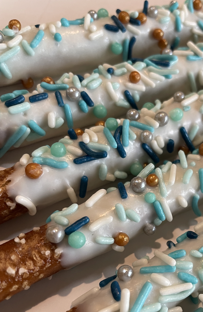 Close-up of pretzel sticks coated in white icing and decorated with blue, white, gold, silver, and light green sprinkles.
