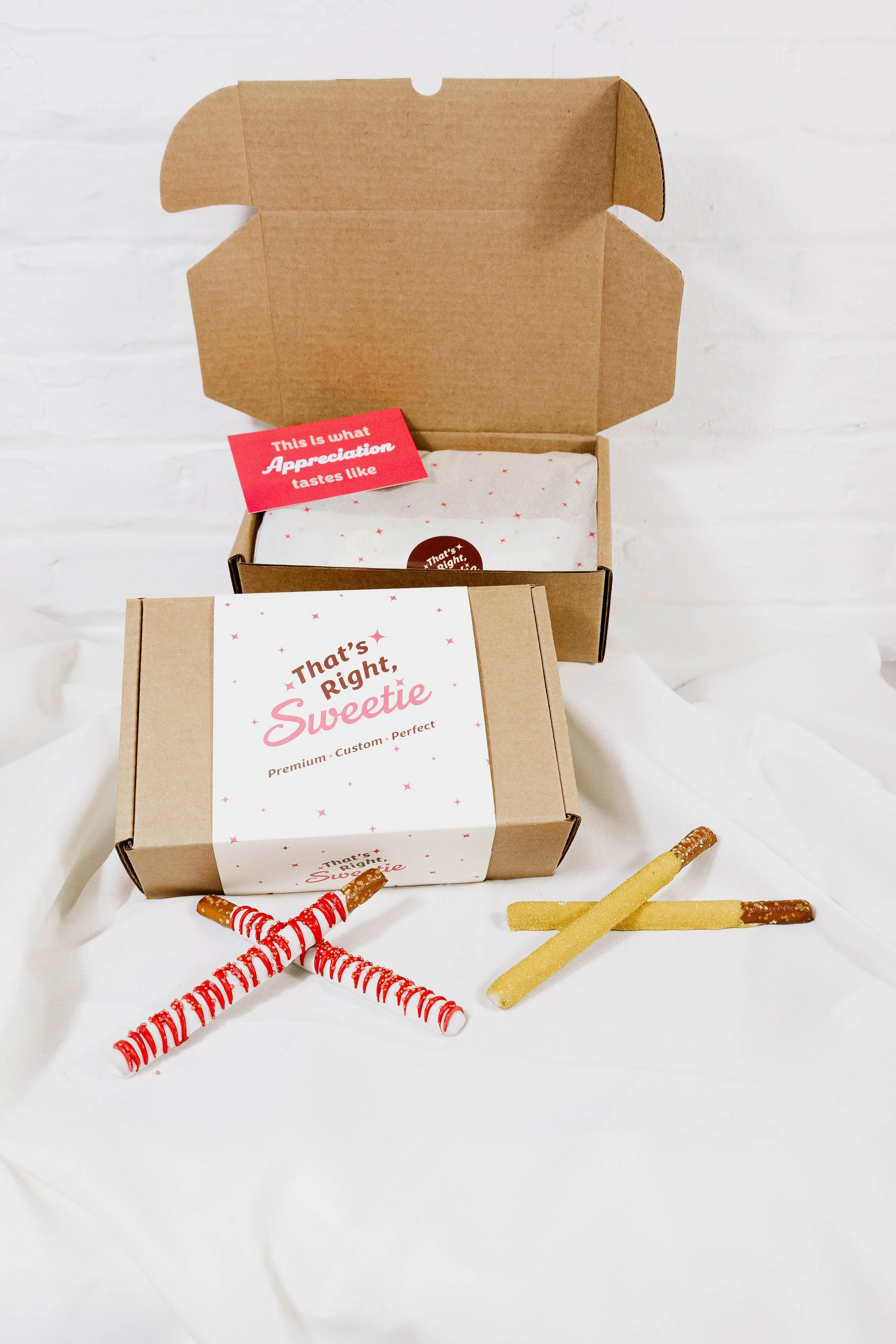 Open brown cardboard gift box with white tissue paper inside, labeled 'That's right, Sweetie,' on a white fabric surface with candy canes, and sugar-coated sour sticks nearby.