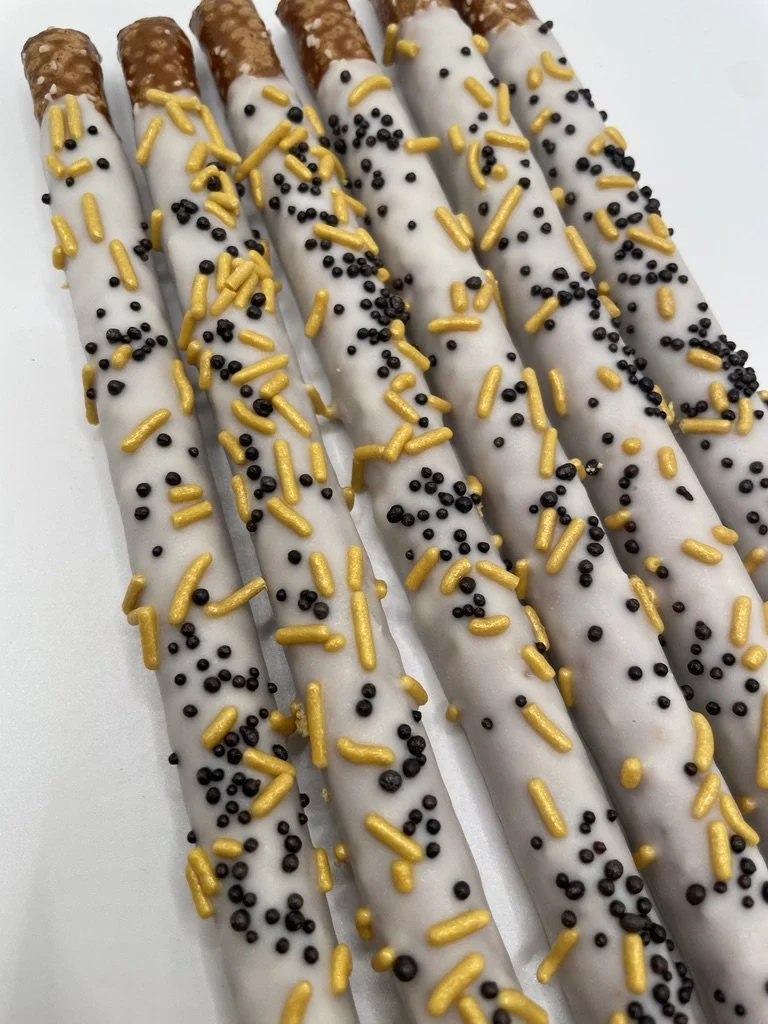 Six pretzel rods coated in white chocolate, decorated with yellow sprinkles and small black round sprinkles.