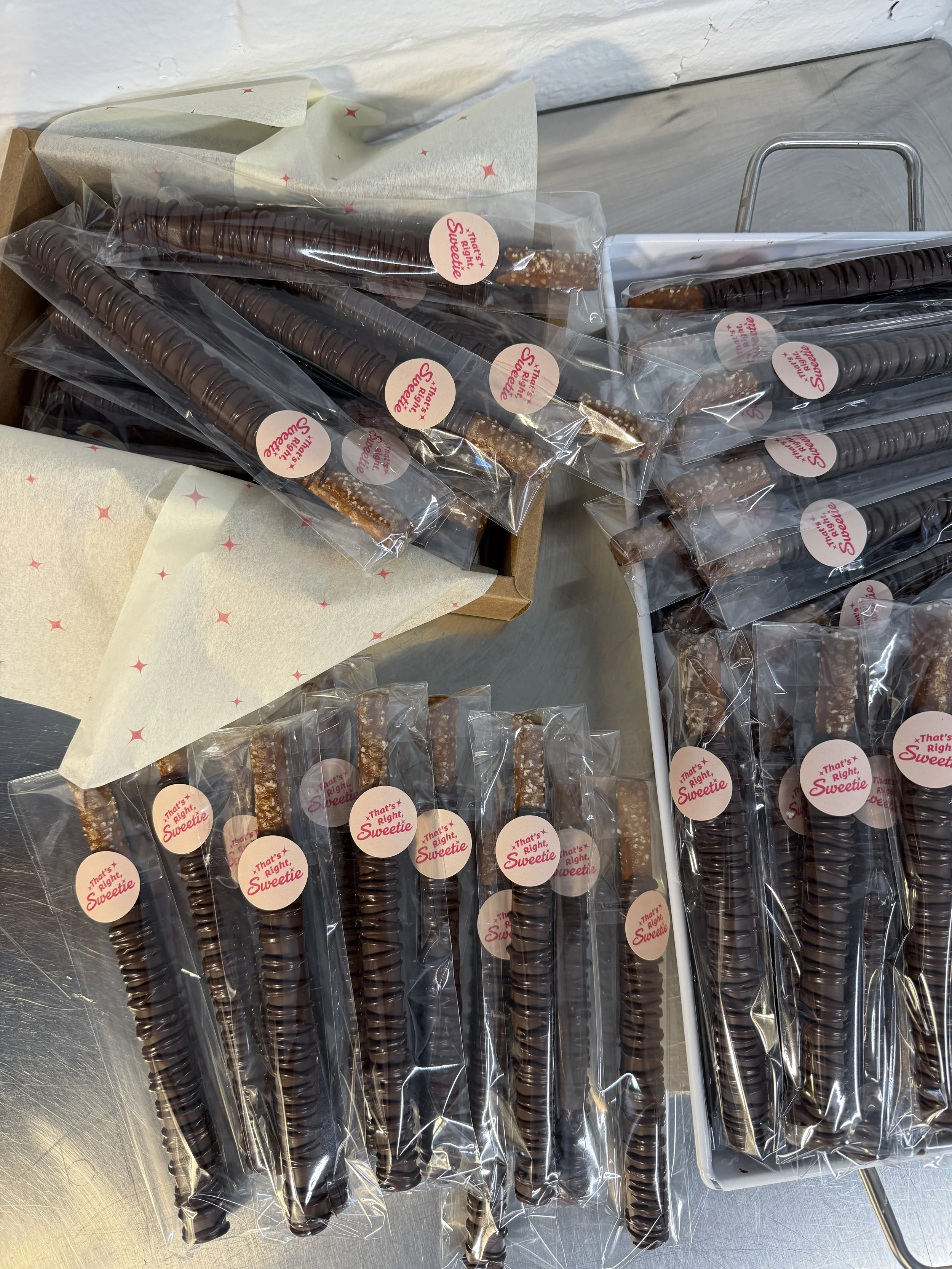 Multiple chocolate-covered pretzel sticks in individual plastic wrappers with a pink sticker that reads 'That's Right, Sweetie' on a metal surface, some wrapped in tissue paper with red star patterns.