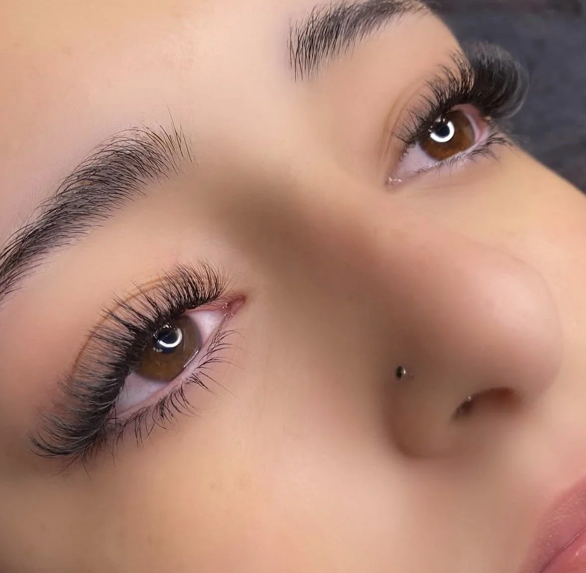 Close-up of a person's face showing brown eyes with long eyelashes, well-groomed eyebrows, and a nose piercing.