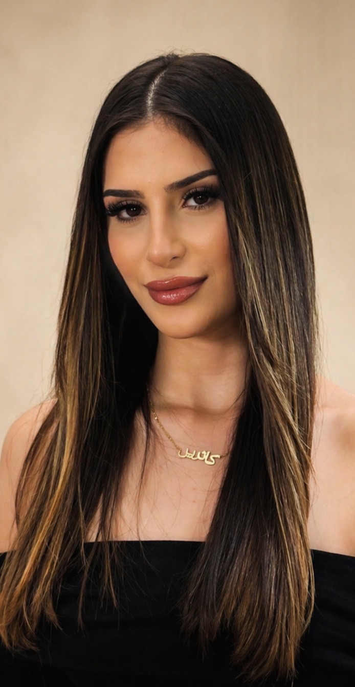 A woman with long straight brown hair and highlights, smiling, wearing makeup with bold lashes and lipstick, a black off-the-shoulder top, and a gold necklace with a name pendant.