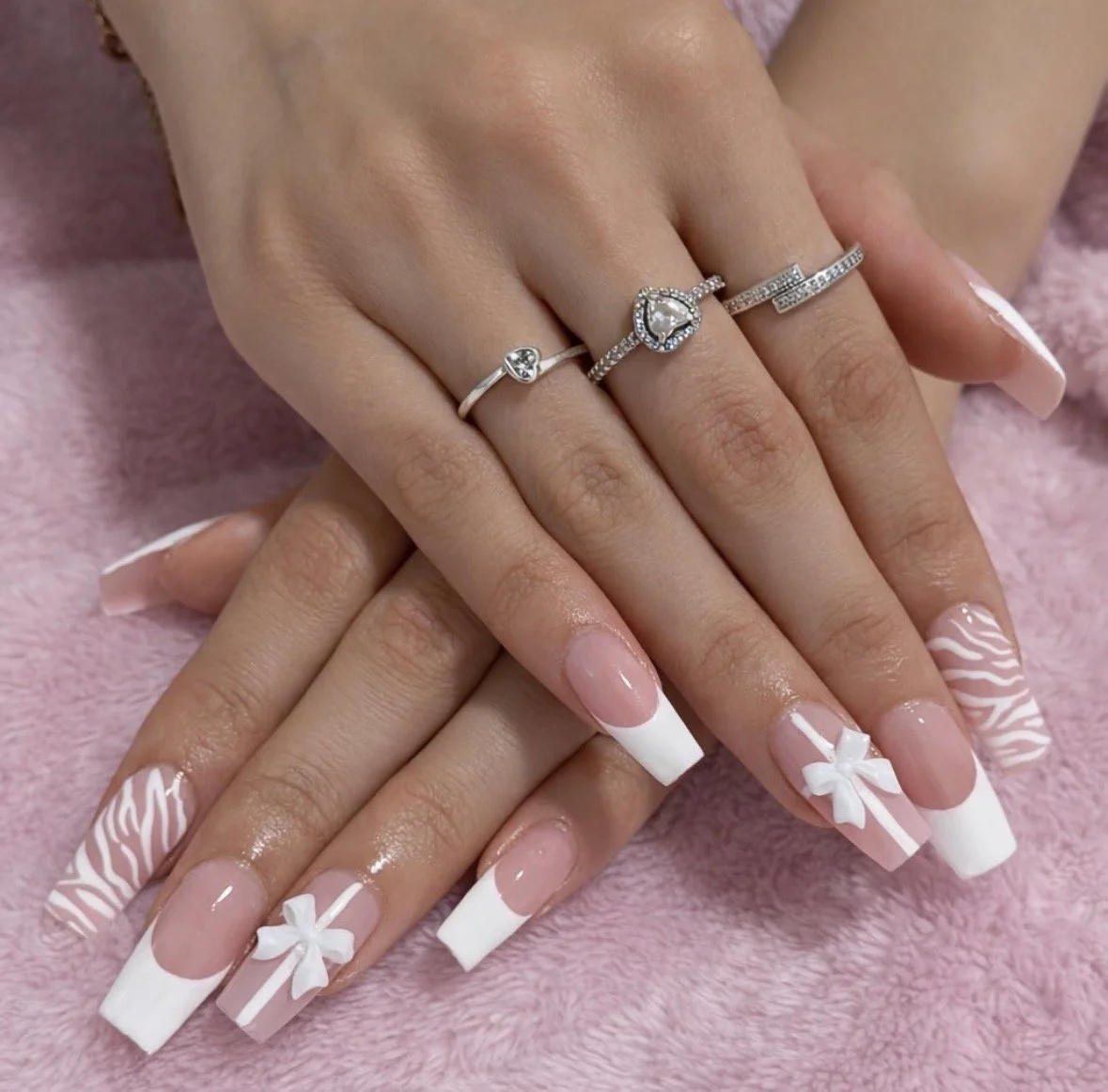 Close-up of hands with manicured nails featuring zebra stripe, pink with a bow, and French tip designs, wearing multiple silver rings with gemstones, resting on a soft pink surface.